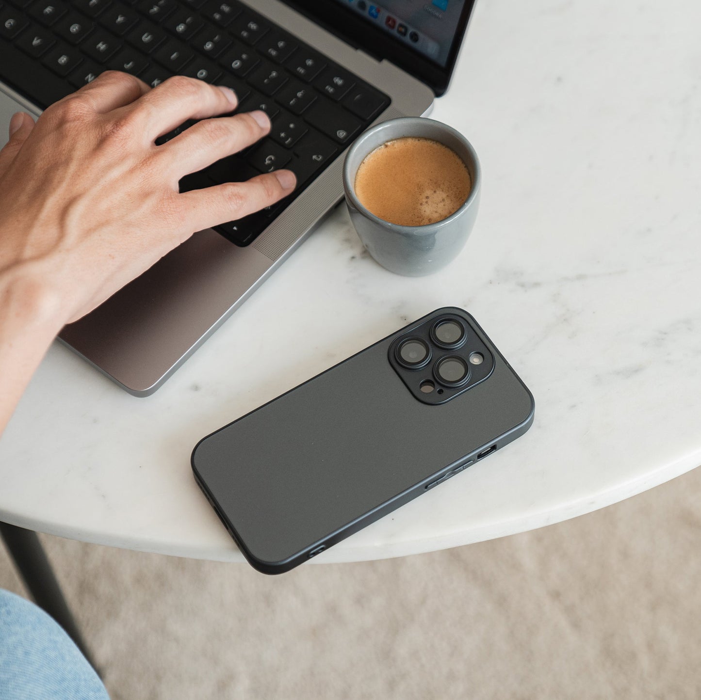 A person types on a laptop at a round white table with a cup of coffee and a smartphone in an arktis iPhone 16e Tech Case with camera protection and AG glass placed nearby.