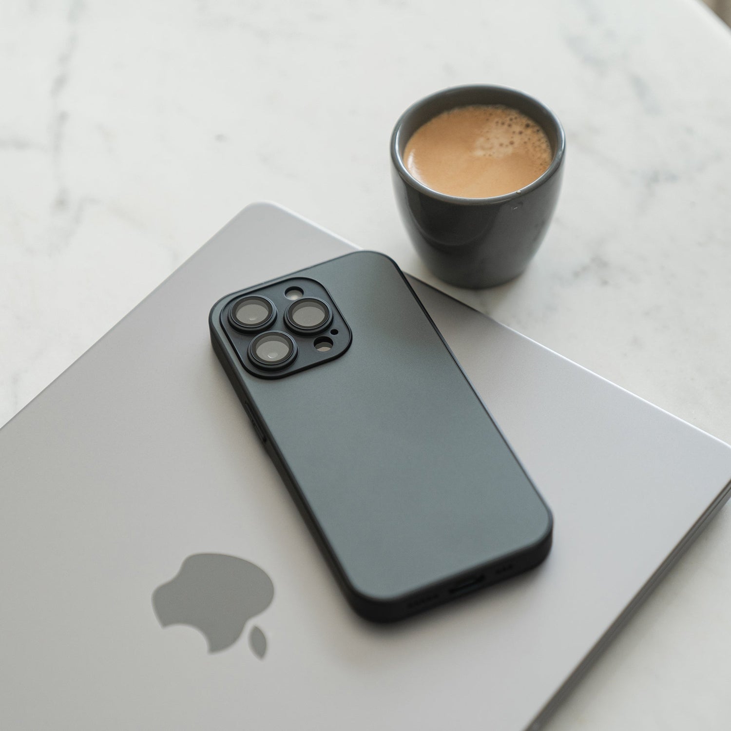 A black smartphone with the arktis iPhone 16e Tech Case lies on a closed silver Apple laptop, next to a small cup of coffee on a white marble surface.