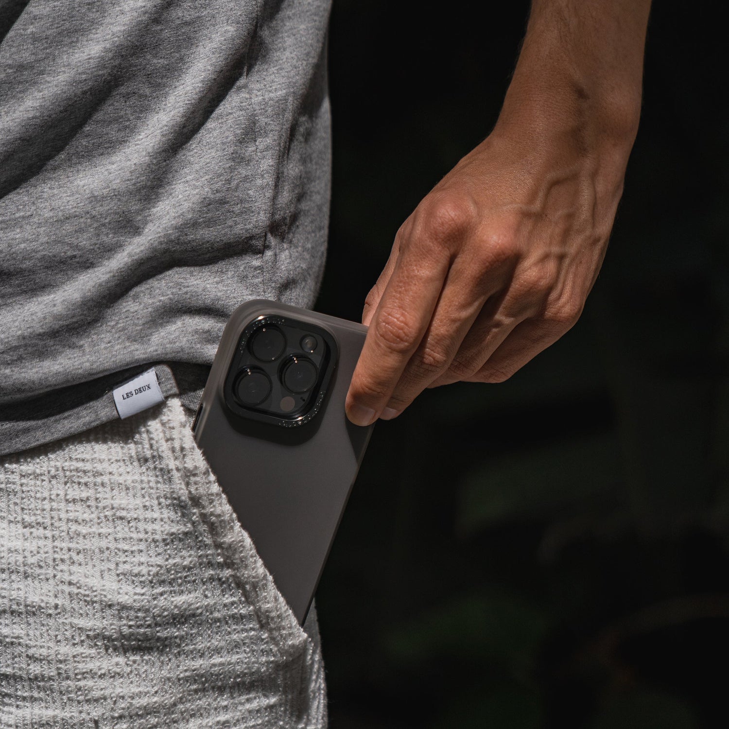 Eine Person steckt ein iPhone 15 Plus mit drei Kameras, geschützt durch das MR PROTECT iPhone 15 Plus FROZEN Case, in die Tasche einer strukturierten Shorts.
