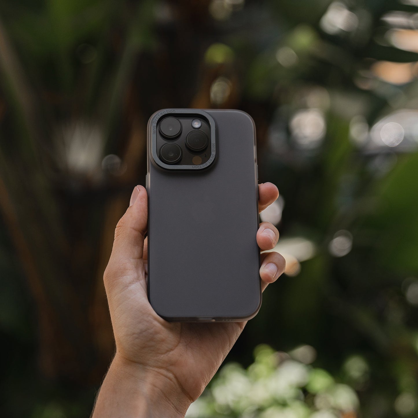 Eine Hand hält das MR PROTECT iPhone 15 Plus FROZEN Case, ein graues Polycarbonat-Case von MR PROTECT, dessen durchscheinendes Aussehen vor verschwommenen grünen Pflanzen im Hintergrund zur Geltung kommt.
