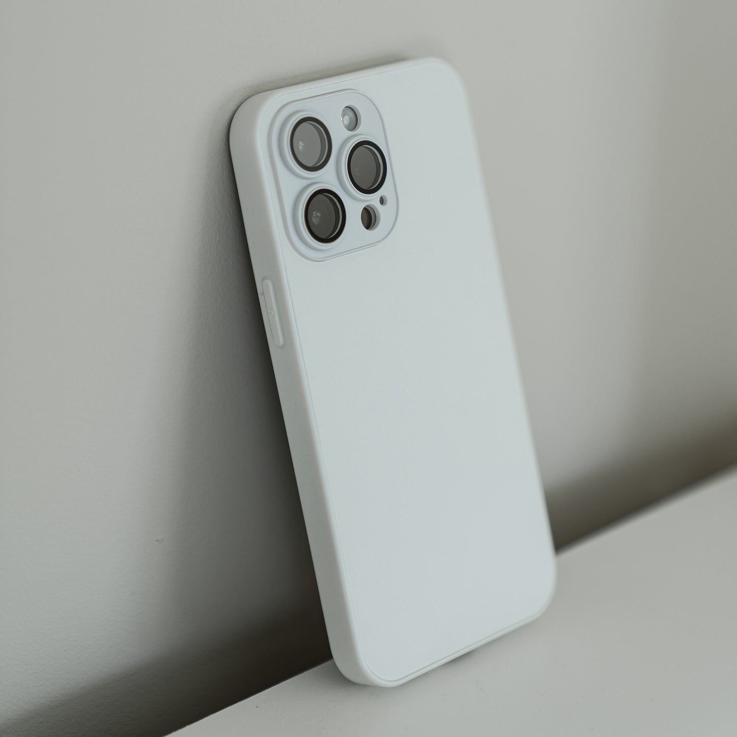 Ein iPhone 15 Plus mit Dreifachkameras und einem schlichten weißen MR PROTECT INVICTUS Case lehnt auf einer ebenen Fläche an einer hellen Wand.