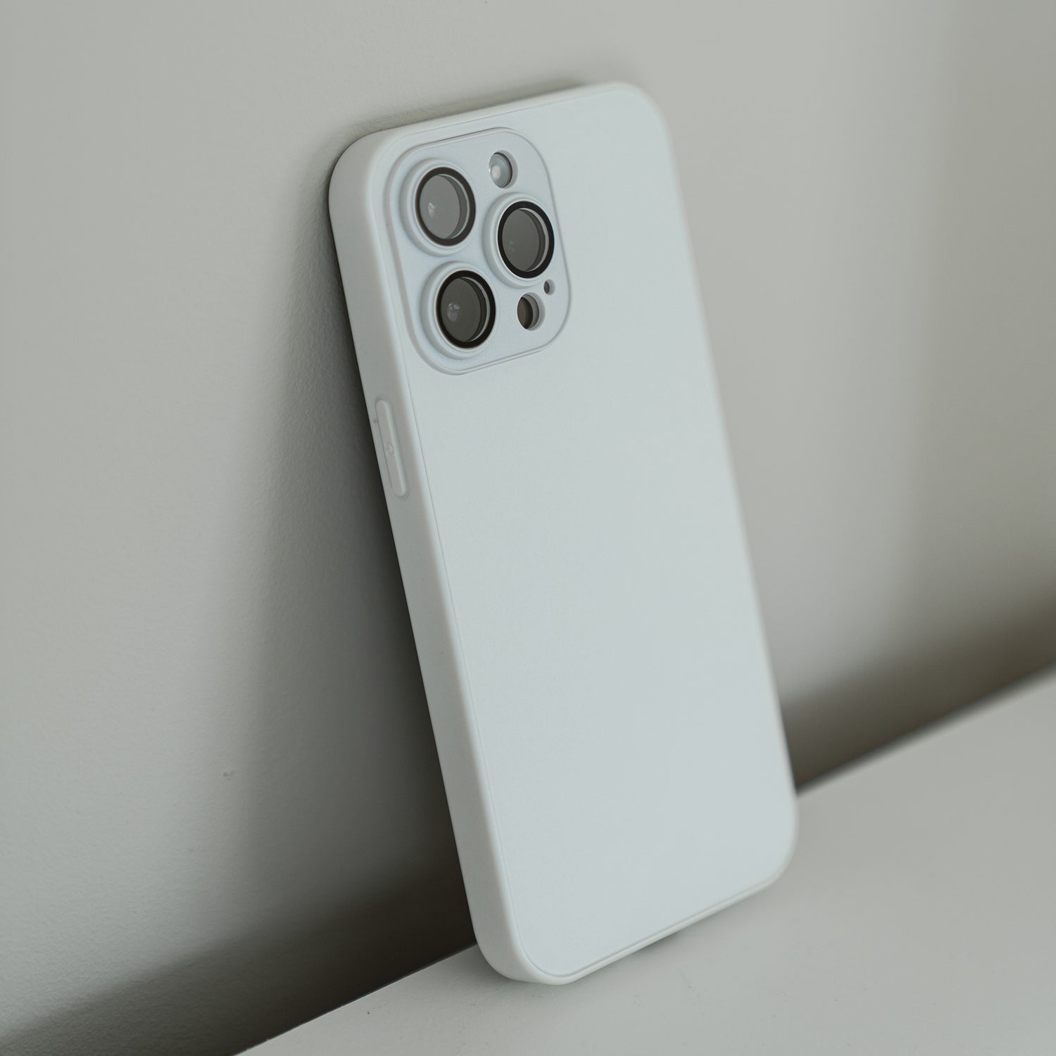 Ein iPhone 15 Plus mit Dreifachkameras und einem schlichten weißen MR PROTECT INVICTUS Case lehnt auf einer ebenen Fläche an einer hellen Wand.