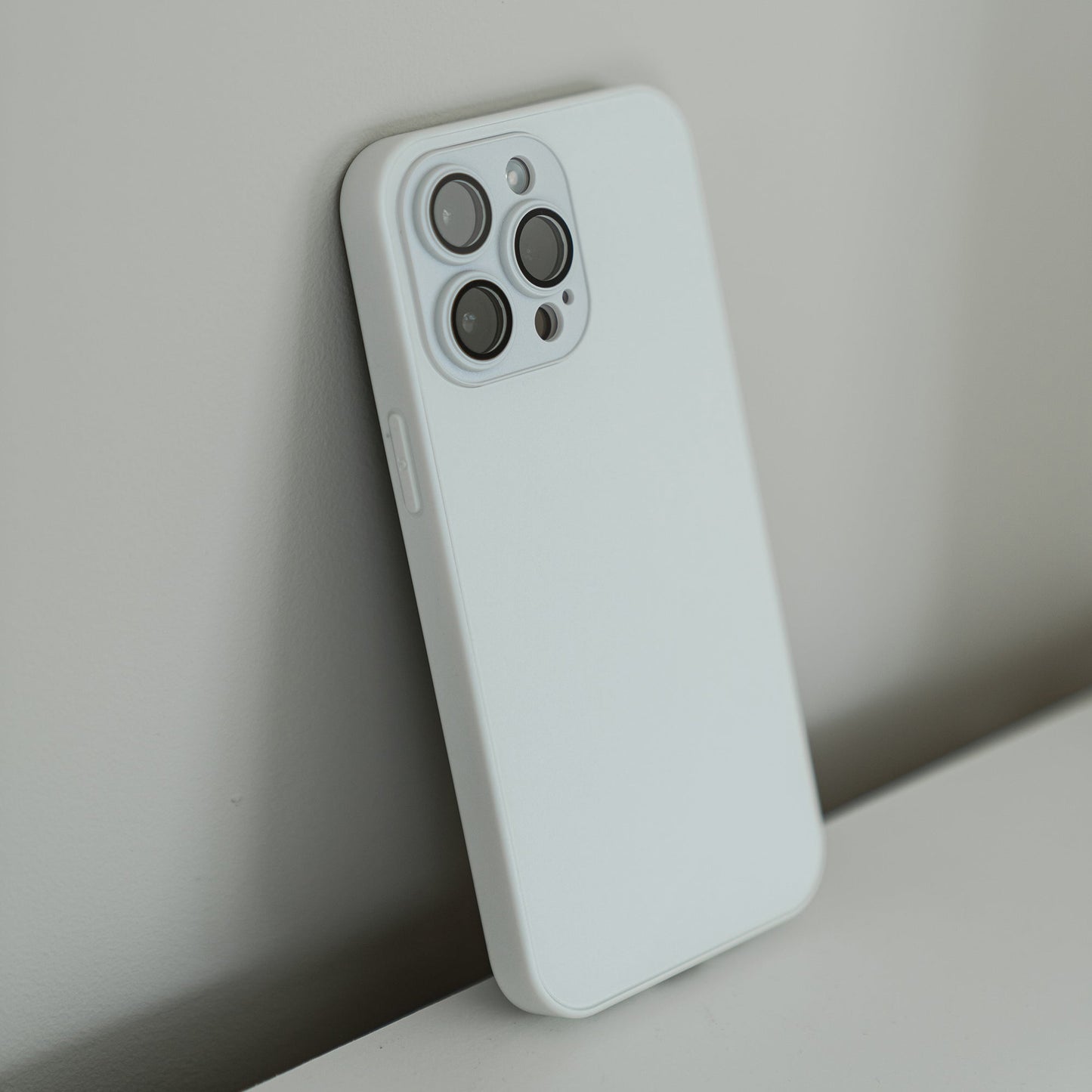 Ein arktis iPhone 16 Tech Case in Weiß aus stoßdämpfendem TPU-Silikon schützt das Smartphone, das schräg an einer hellen Wand oder Oberfläche lehnt. Die Kameralinsen und der AG-Glas-Blitz sind auf der Rückseite gut sichtbar.