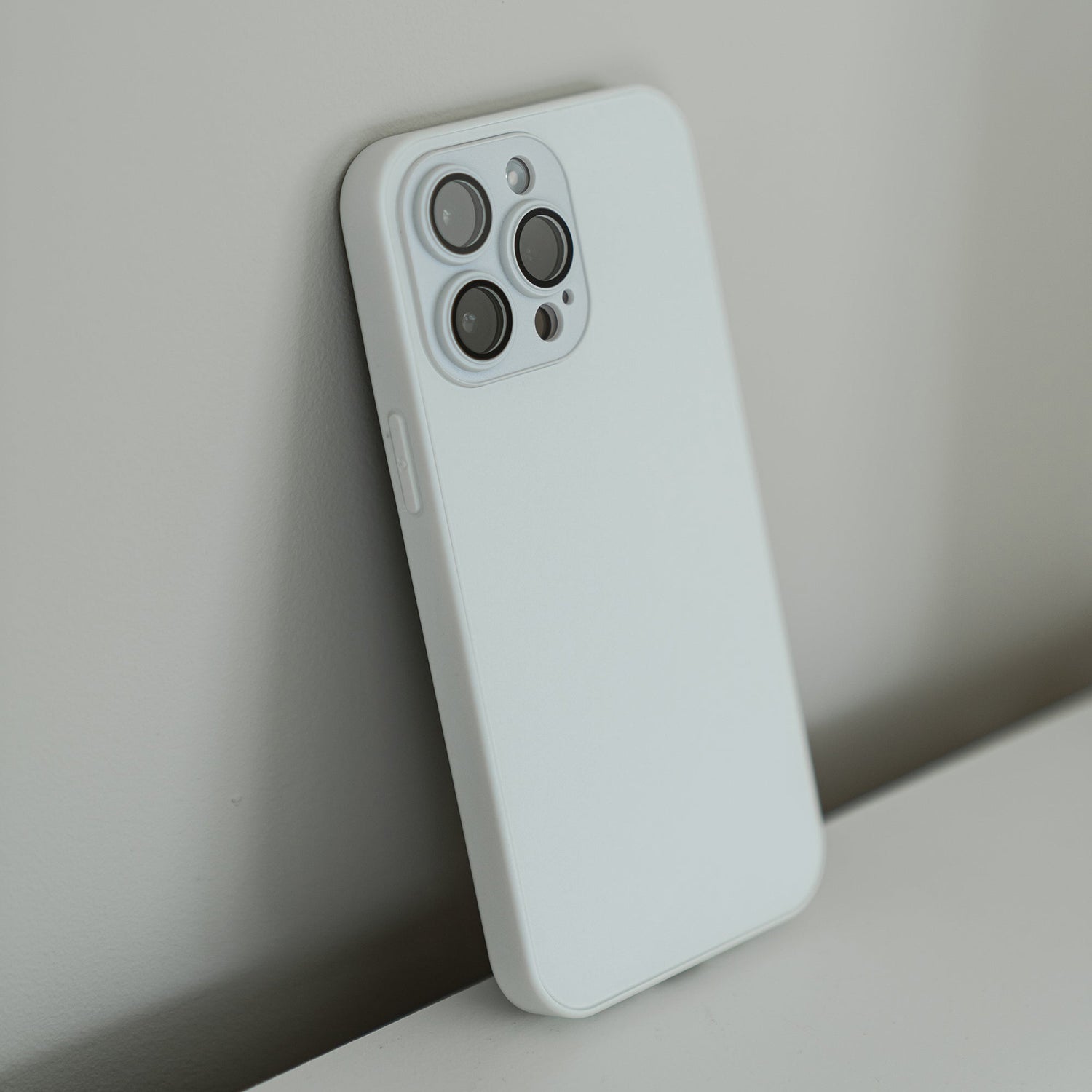 Ein arktis iPhone 16 Tech Case in Weiß aus stoßdämpfendem TPU-Silikon schützt das Smartphone, das schräg an einer hellen Wand oder Oberfläche lehnt. Die Kameralinsen und der AG-Glas-Blitz sind auf der Rückseite gut sichtbar.