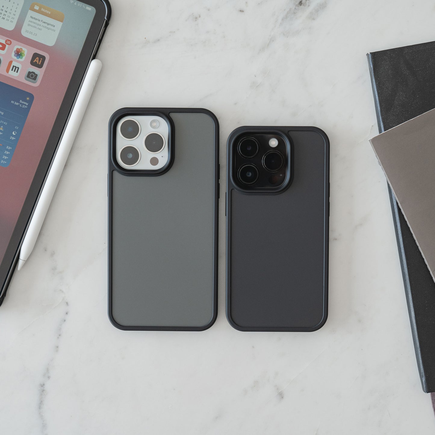 Zwei MR PROTECT iPhone 15 Case Matt von MR PROTECT mit Stoßabsorption liegen nebeneinander auf weißem Marmor, daneben ein Tablet mit Stylus und ein Stapel geschlossener Notizbücher im minimalistischen Design.