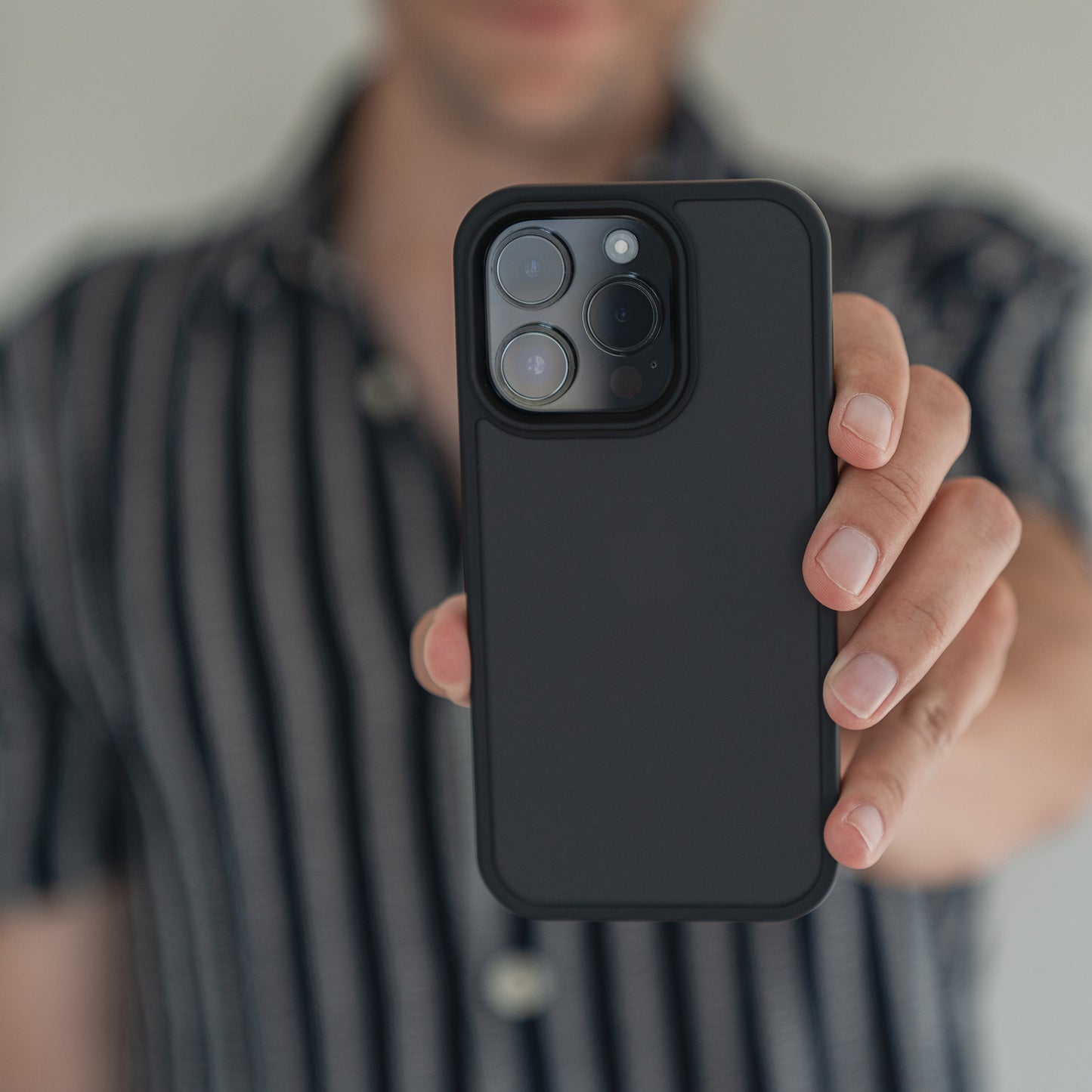 Eine Person in einem schwarz-weiß gestreiften Hemd hält das MR PROTECT iPhone 15 Pro Case Matt mit stoßabsorbierendem TPU-Silikon und minimalistischem Matt-Look in Richtung Kamera und verdeckt so teilweise ihr Gesicht. Die Dreifachkamera des Handys ist sichtbar.