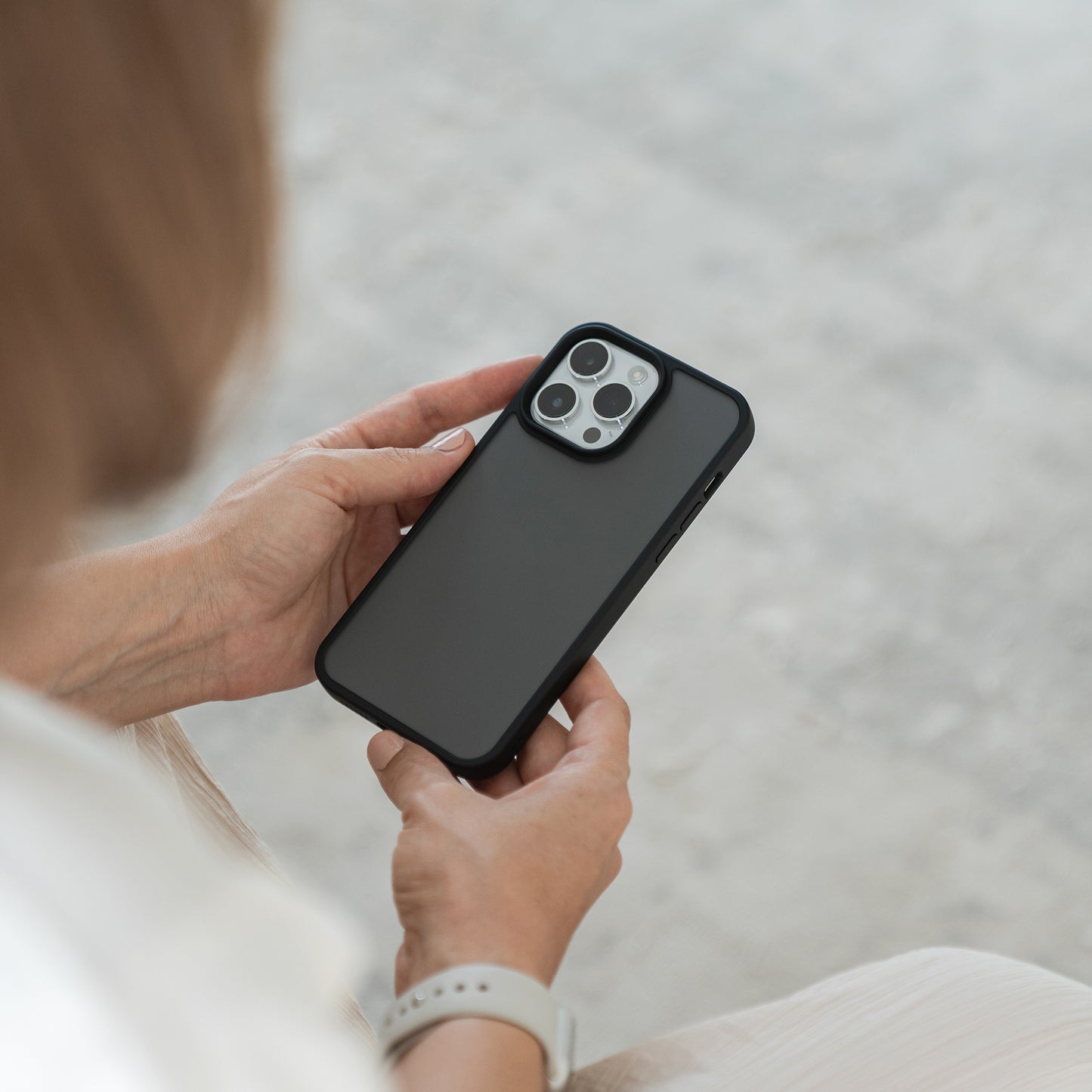 Eine Person hält ein MR PROTECT iPhone 15 Case Matt von MR PROTECT mit beiden Händen. Das Telefon liegt mit der Vorderseite nach unten, sodass die Kameralinsen sichtbar sind. Sie trägt ein helles Hemd und eine Smartwatch, während der Hintergrund unscharf bleibt.