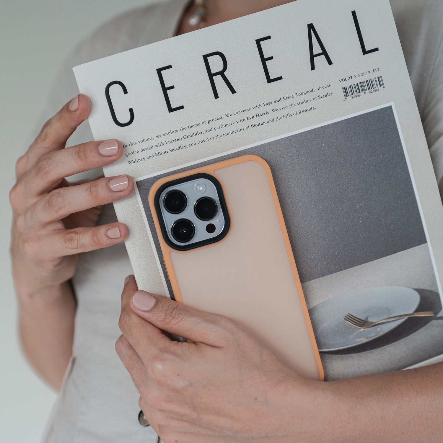 Eine Person, vom Oberkörper abwärts gesehen, hält ein Cereal-Magazin und ein Smartphone mit dem MR PROTECT iPhone 15 Pro Case Matt von MR PROTECT; ihre Nägel sind in einem hellen Farbton manikürt.
