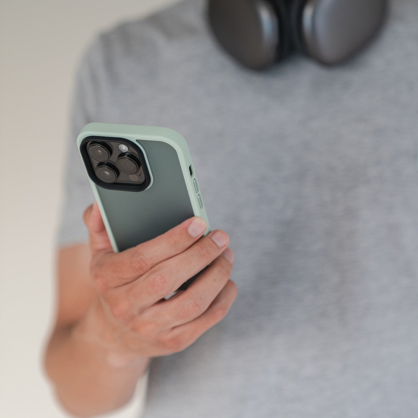 Eine Person mit grauem T-Shirt und Kopfhörern hält ein Smartphone mit dem MR PROTECT iPhone 15 Case Matt in Grün. Das minimalistische Case von MR PROTECT lässt die Kameralinsen des Handys gut sichtbar.