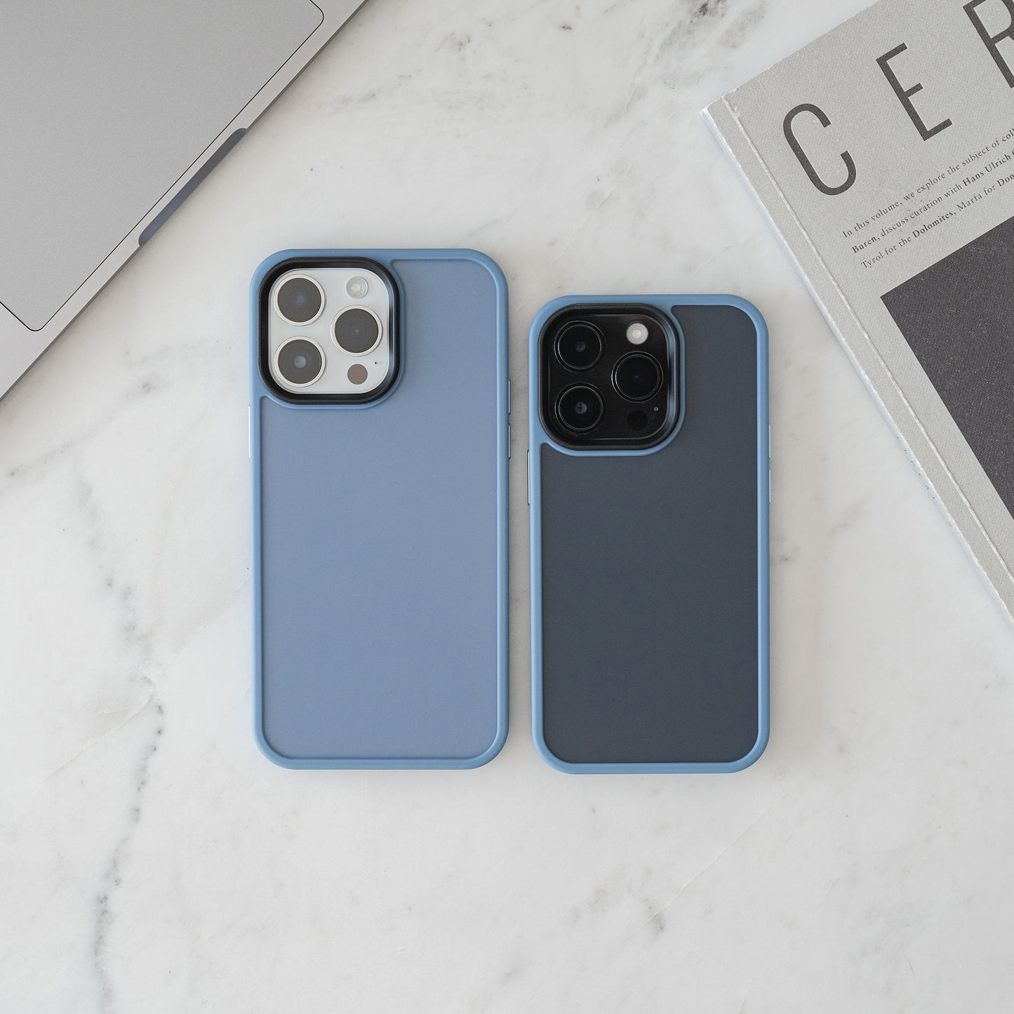 Zwei MR PROTECT iPhone 15 Case Matt Hüllen in Blau aus stoßabsorbierendem TPU-Silikon liegen im minimalistischen Look nebeneinander auf weißem Marmor, neben einem Laptop-Ausschnitt und einer Zeitschrift.