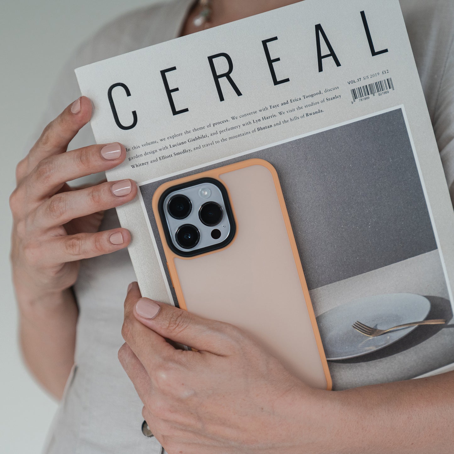Eine Person hält das Cereal-Magazin und eine MR PROTECT iPhone 15 Hülle Matt in der Hand. Das Magazin zeigt einen minimalistischen Teller, eine Gabel und einen Löffel auf einem grauen Cover. Ihre Nägel sind hellrosa lackiert.