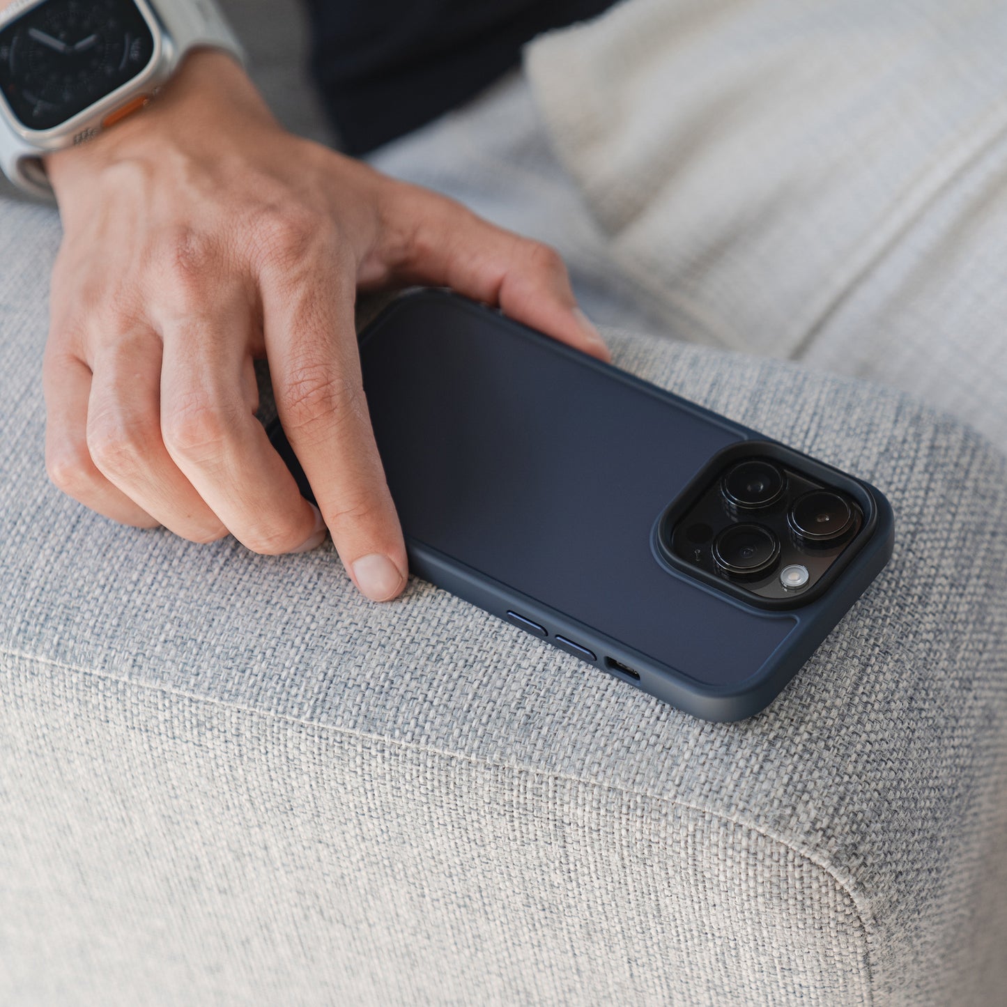 Die Hand einer Person mit einer Smartwatch ruht auf einer hellgrauen Stoffoberfläche neben einem mit der Vorderseite nach unten gerichteten MR PROTECT iPhone 15 Case Matt von MR PROTECT in minimalistischem Schwarz.