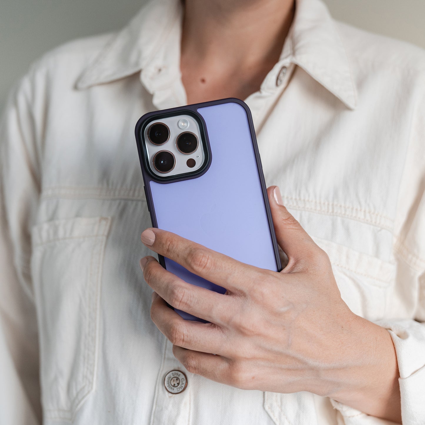 Eine Person in einem hellen Hemd mit Knöpfen hält ein iPhone 15 mit dem MR PROTECT iPhone 15 Case Matt von MR PROTECT mit minimalistischem Matt-Look und dreifacher Kameralinse nahe ihrer Brust.