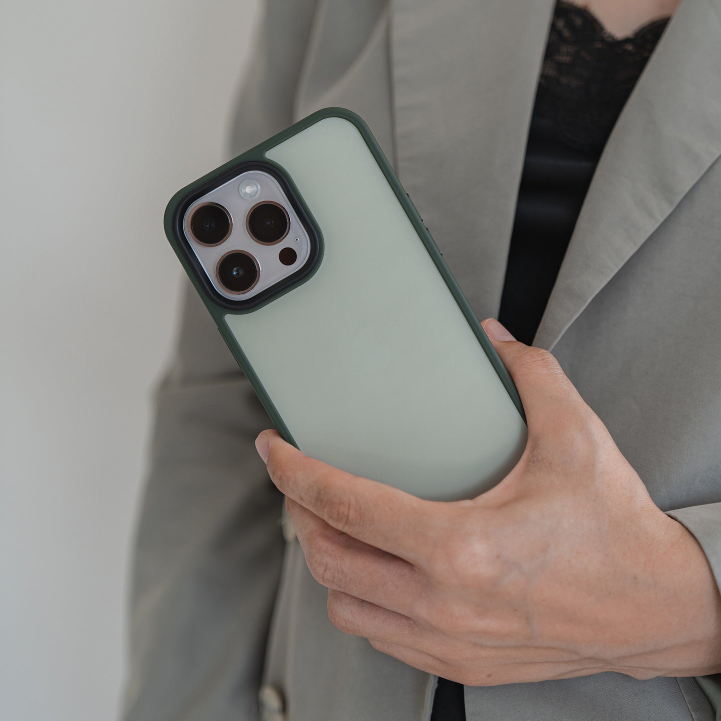 Eine Person in grauem Blazer und schwarzem Spitzentop hält das MR PROTECT iPhone 15 Case Matt im grünen Minimalist-Look in der rechten Hand. Der Fokus liegt auf Handyhülle und Hand, ein Teil des Blazers ist sichtbar.