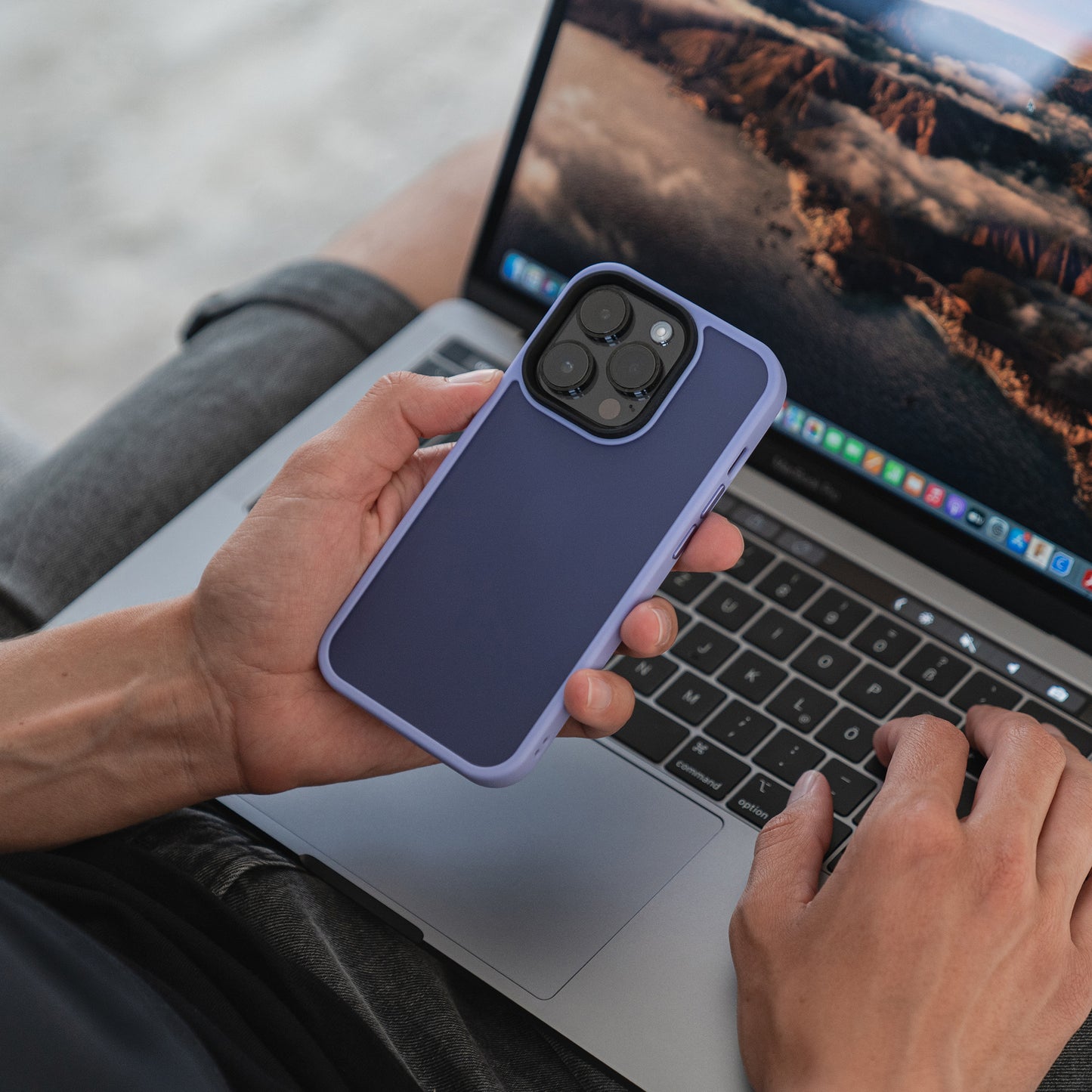 Eine Person hält mit einer Hand ein MR PROTECT iPhone 15 Case Matt in Blau mit Stoßdämpfung und minimalistischem Matt-Look, während sie auf einem Laptop tippt, auf dem ein Naturlandschafts-Hintergrundbild angezeigt wird.