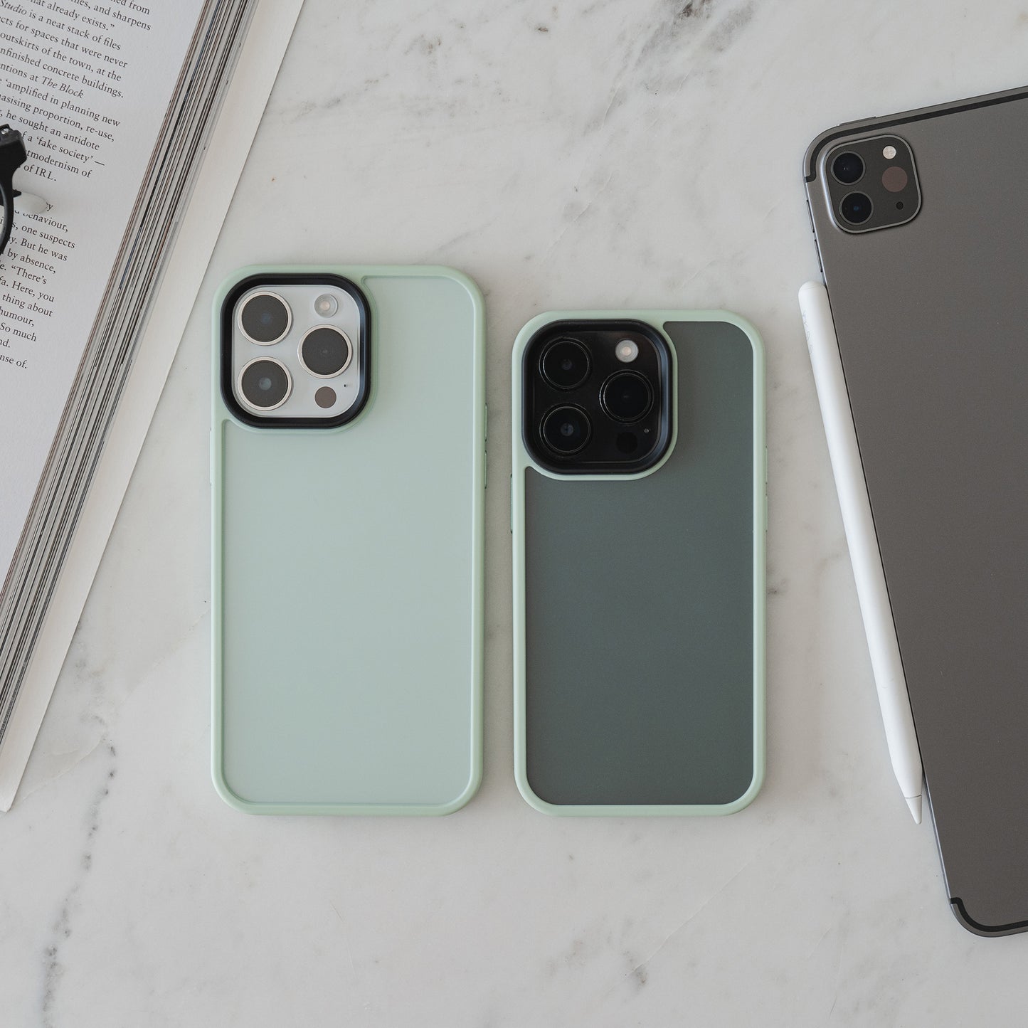Zwei Smartphones mit der matt-minimalistischen MR PROTECT iPhone 15 Pro Case Matt von MR PROTECT liegen nebeneinander auf Marmor, neben einem geschlossenen Tablet mit Stylus und einem geöffneten Buch oder Magazin.