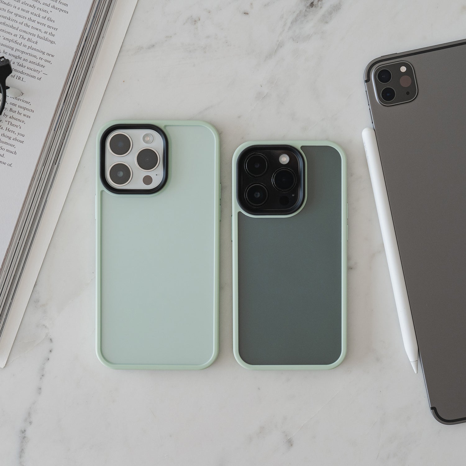 Zwei Smartphones mit der matt-minimalistischen MR PROTECT iPhone 15 Pro Case Matt von MR PROTECT liegen nebeneinander auf Marmor, neben einem geschlossenen Tablet mit Stylus und einem geöffneten Buch oder Magazin.