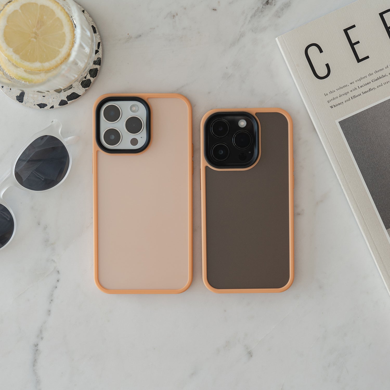 Zwei Smartphones im orange umrandeten MR PROTECT iPhone 15 Pro Case Matt von MR PROTECT mit stoßdämpfendem TPU-Silikon liegen auf Marmor neben einem Glas Zitronenwasser, einer weißen Sonnenbrille und einem CER-Magazin.