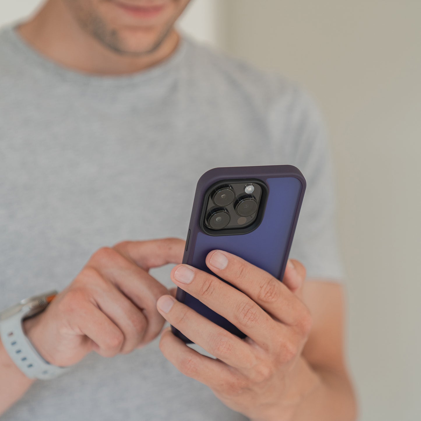 Eine Person in hellgrauem T-Shirt und Smartwatch hält das MR PROTECT iPhone 15 Case Matt mit minimalistisch-mattem Blau-Finish und tippt mit der rechten Hand auf den Bildschirm. Der Hintergrund ist unscharf.