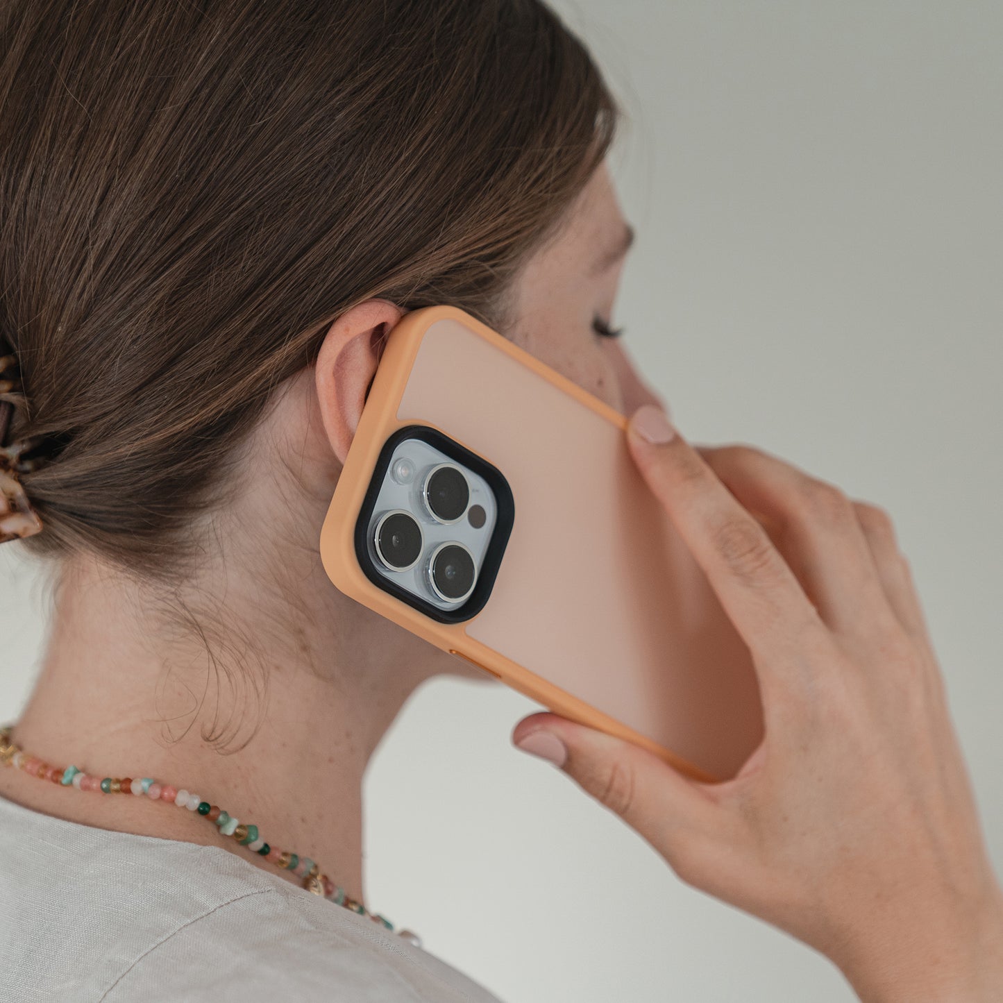 Eine Person mit braunem Haarknoten hält ein MR PROTECT iPhone 15 Case Matt Peach-Smartphone mit Dreifachkamera ans Ohr, als würde sie telefonieren. Sie trägt eine Perlenkette und ein helles Oberteil.