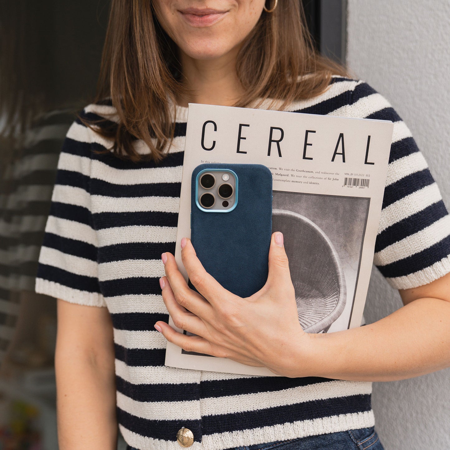 Eine Frau im gestreiften Hemd steht an einer Wand, hält eine Zeitschrift in der Hand und ihr Handy geschützt durch die arktis iPhone Air Hülle aus Alcantara von arktis.