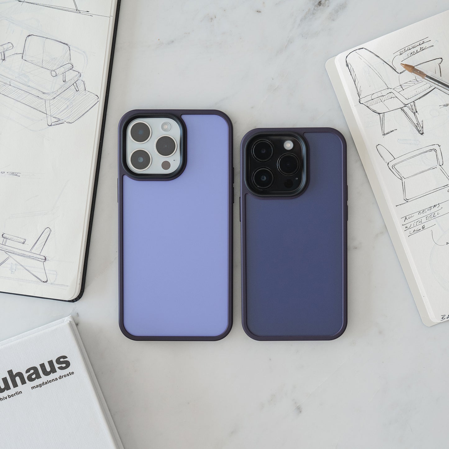 Zwei Smartphones mit MR PROTECT iPhone 15 Case Matt von MR PROTECT liegen auf einer weißen Fläche neben offenen Skizzenbüchern mit Stuhlentwürfen, einem Bauhaus-Buch und einem Bleistift, der auf einem Skizzenbuch liegt.