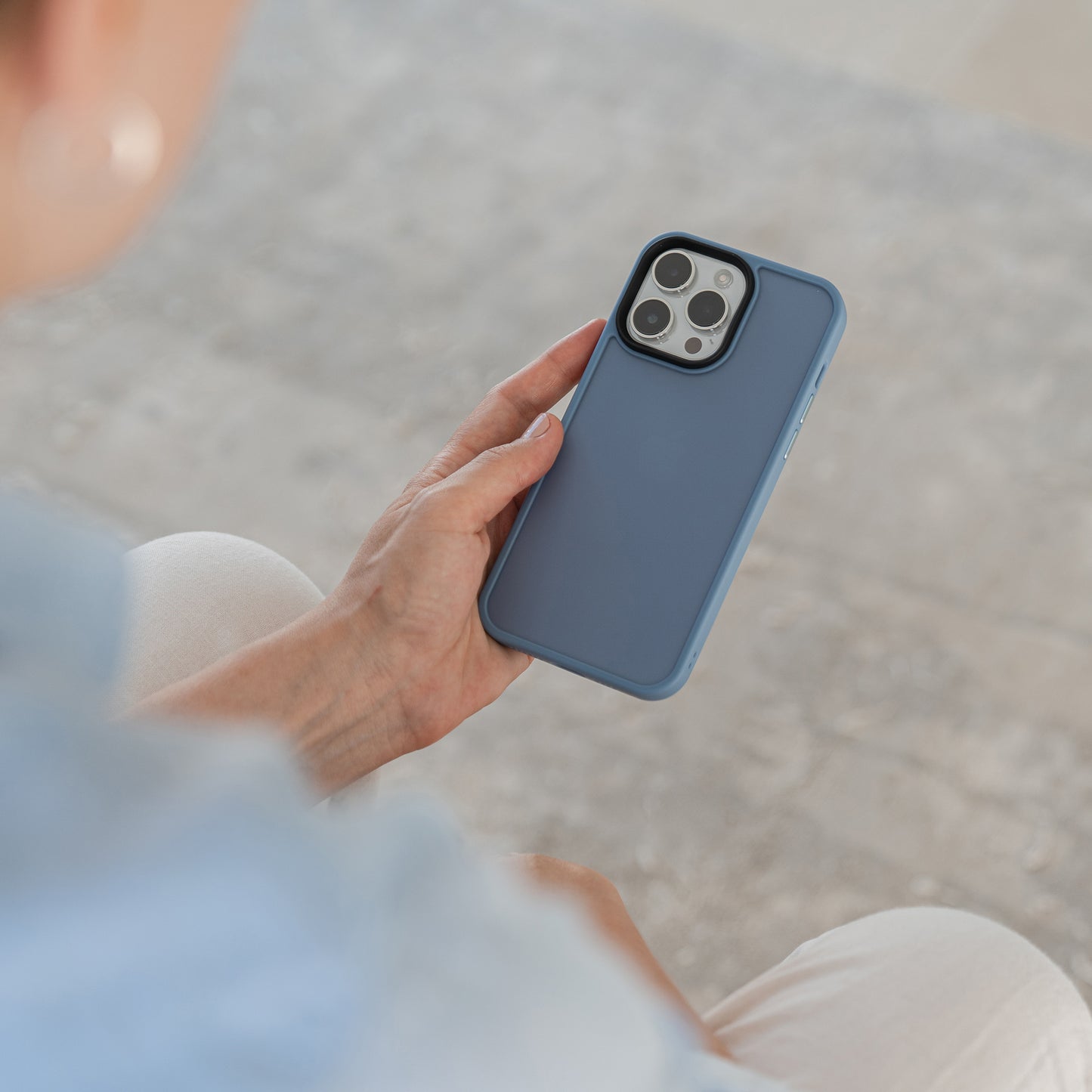Eine Person hält ein iPhone 15 mit drei Rückkameras im MR PROTECT iPhone 15 Case Matt von MR PROTECT in minimalistischer Mattoptik, während sie auf einer hellen Oberfläche sitzt und den Arm auf dem Schoß ruht.