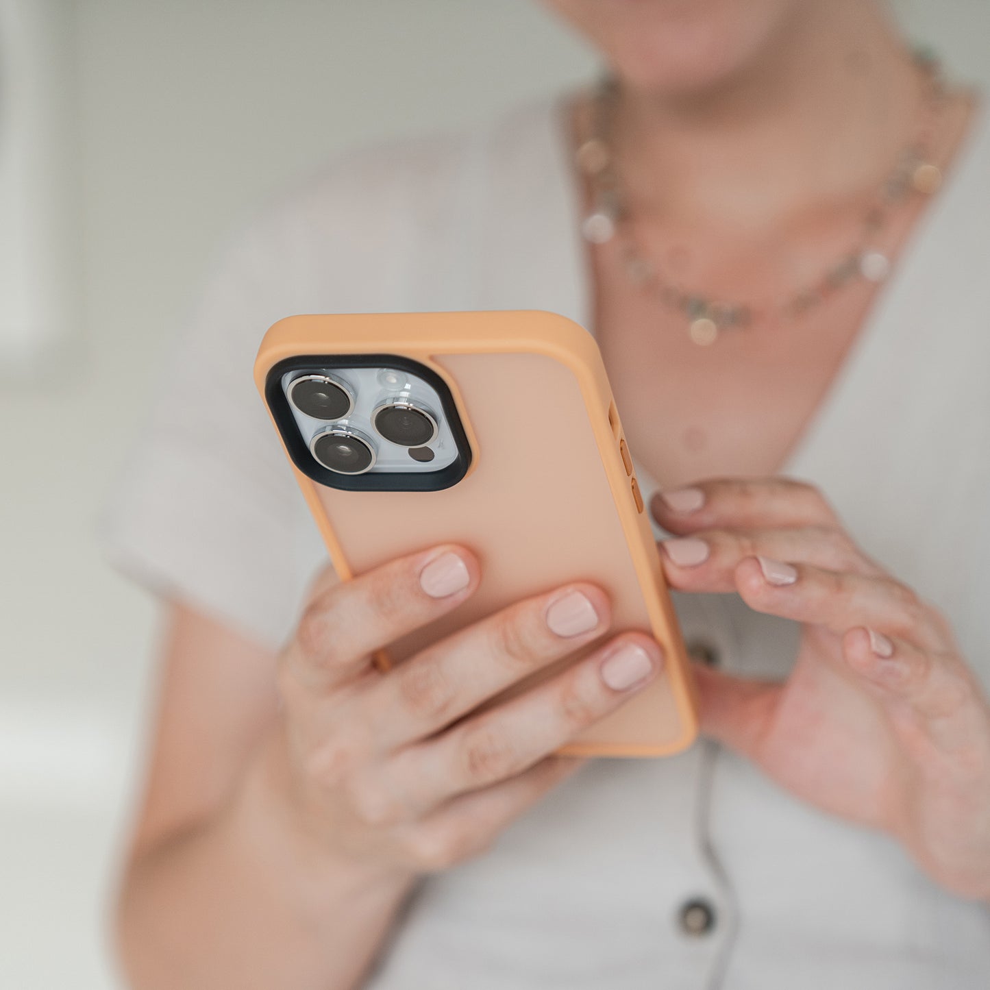 Eine Person in beigem Oberteil und Halskette hält das MR PROTECT iPhone 15 Pro Case Matt, eine minimalistische Hülle in Pfirsichfarbe. Ihre rechte Hand berührt den Bildschirm, wobei der Fokus auf dem Telefon und den Händen liegt, während ihr Gesicht unscharf ist.