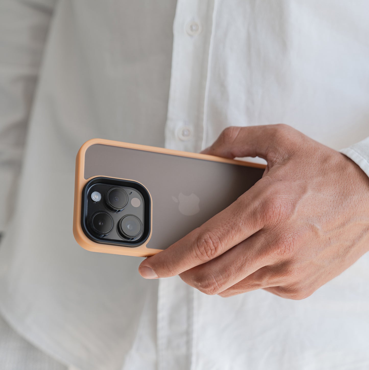 Eine Person in einem weißen Hemd hält das MR PROTECT iPhone 15 Case Matt von MR PROTECT und präsentiert seine braune Farbe mit orangefarbenen Rändern, den minimalistischen matten Look, das sichtbare Apple-Logo und die dreifache Kameraaussparung sowie die zuverlässige Stoßdämpfung.
