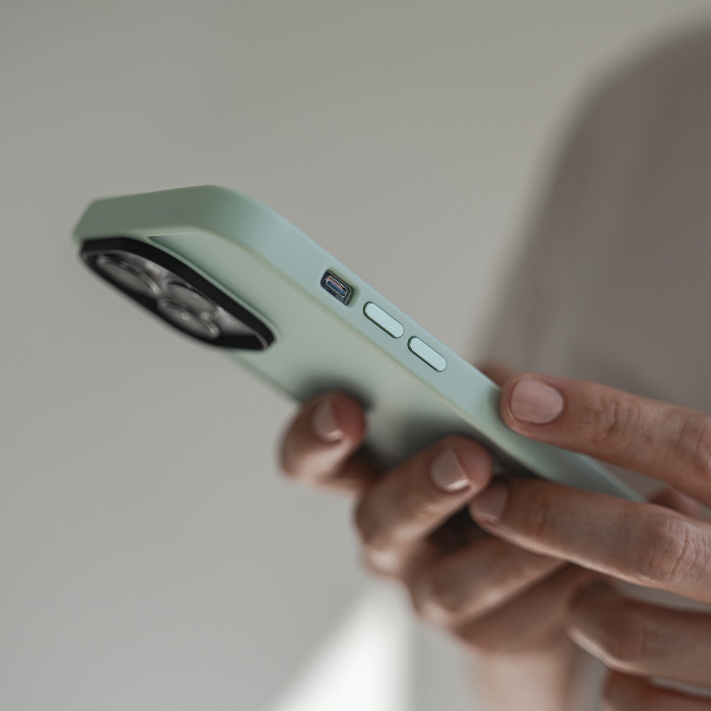 Eine Person hält eine MR PROTECT iPhone 15 Hülle Matt in Hellgrün mit drei sichtbaren Knöpfen, Ladeanschluss und mattem Finish. Kamera-Linie und Hand sind erkennbar, der Hintergrund ist neutral und unscharf.