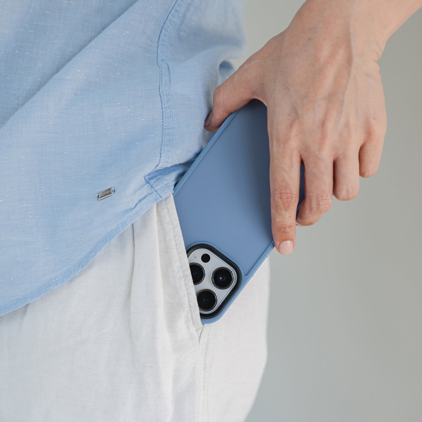 Eine Person in einem hellblauen Hemd und einer weißen Hose steckt ein MR PROTECT iPhone 15 Pro Case Matt in die Tasche, wobei der Fokus auf der Hand liegt, die das MR PROTECT Case hält, und auf dem Taschenbereich.