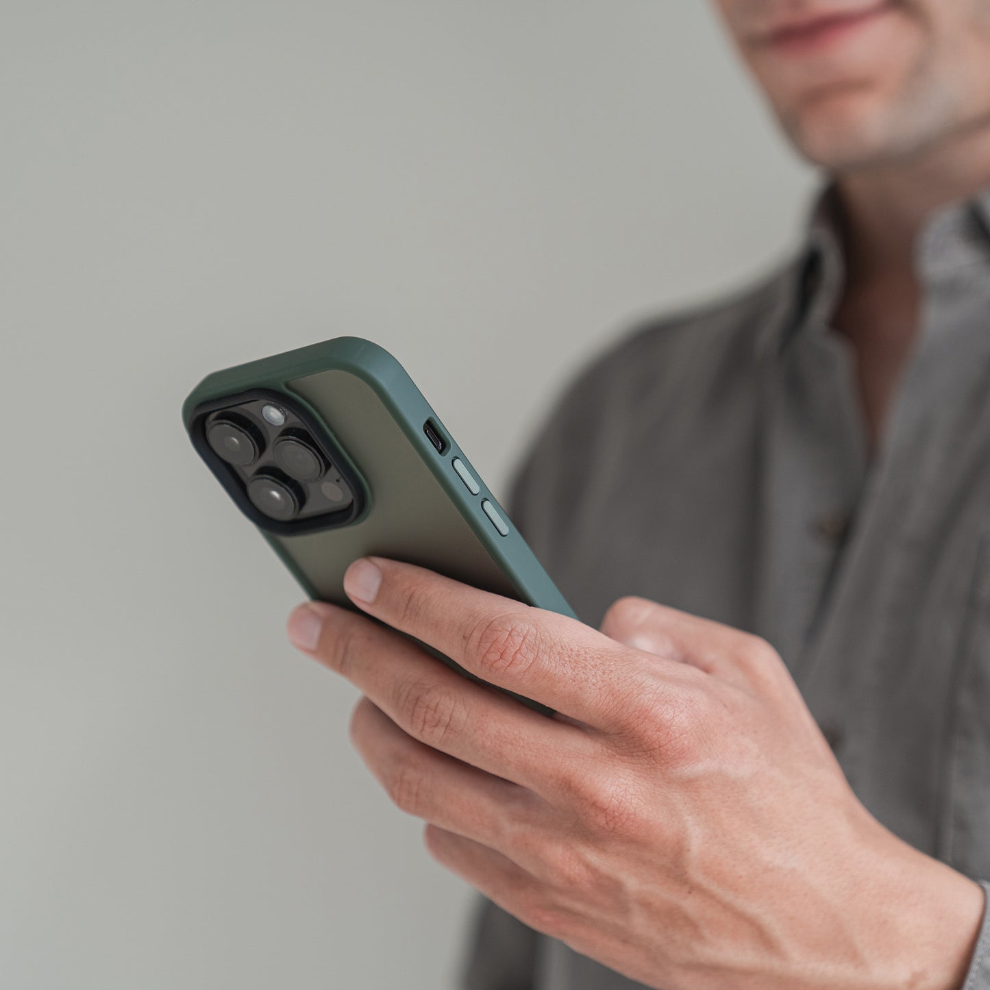 Eine Person in einem grauen Hemd hält ein Smartphone mit dem MR PROTECT iPhone 15 Pro Case Matt von MR PROTECT in Grün und hebt das minimalistische matte Design vor einem schlichten, unscharfen Hintergrund hervor, während der Fokus auf dem Bereich der Rückkamera liegt.