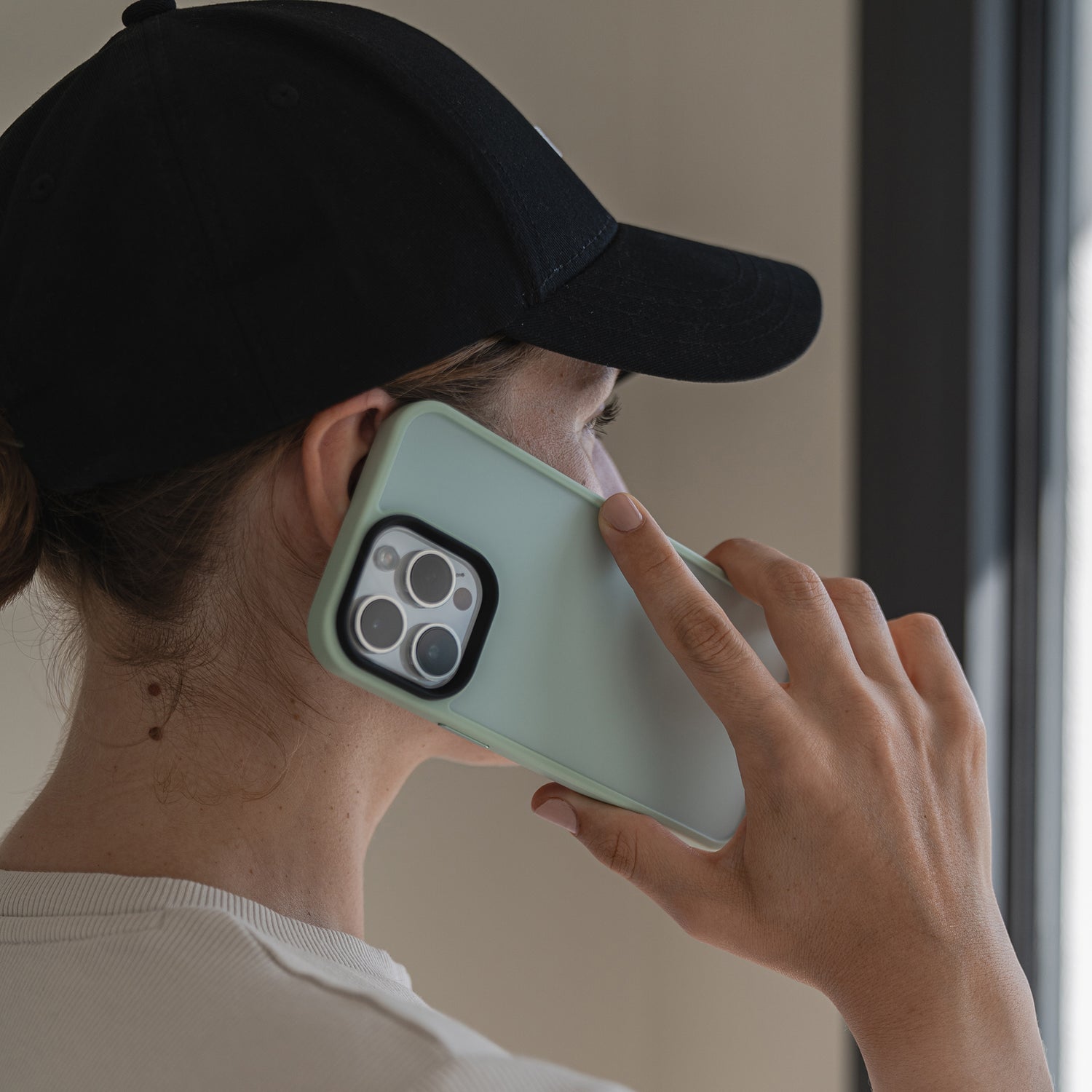 Eine Person mit schwarzer Mütze und weißem Hemd hält ein hellgrünes MR PROTECT iPhone 15 Pro Case Matt ans Ohr und telefoniert in einem Innenbereich an einem Fenster mit Tageslicht.