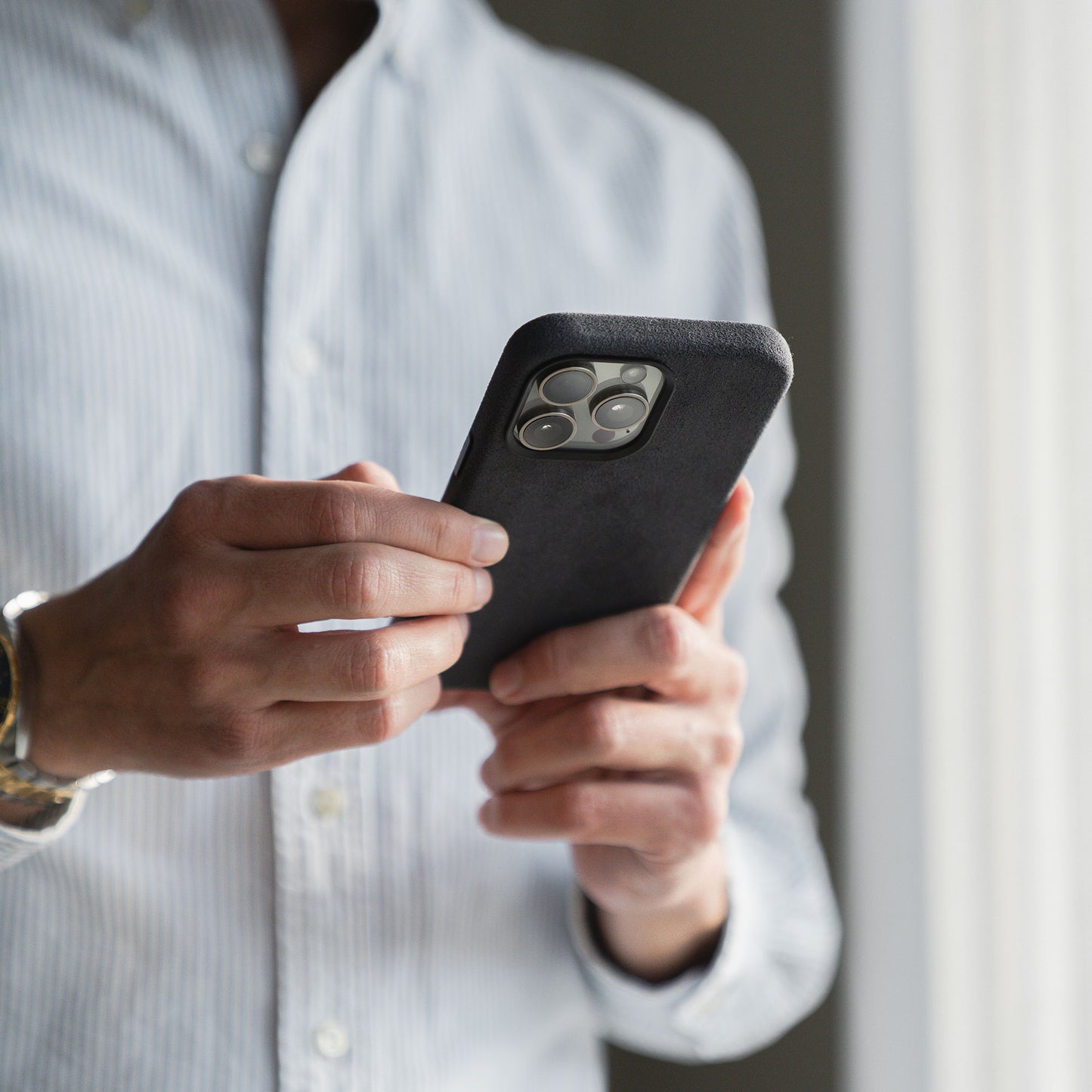 Person im gestreiften Hemd, die ein Smartphone mit einer arktis iPhone Air Hülle aus Alcantara von arktis in der Nähe eines Fensters hält, durch das Licht strömt.