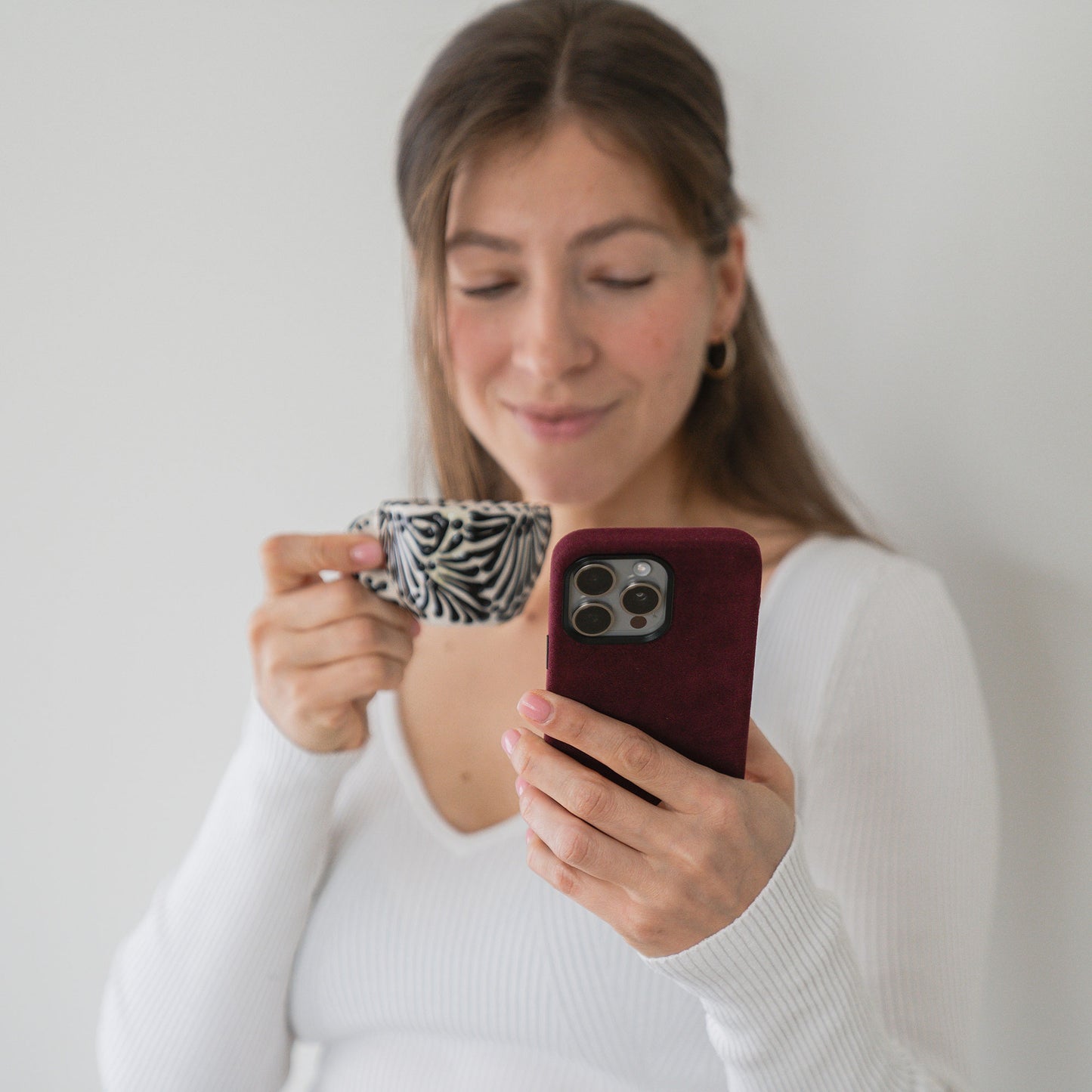 Frau in weißem Oberteil hält eine gemusterte Tasse in der Hand und schaut leicht lächelnd auf ihr Handy, das von der stylischen arktis iPhone Air Hülle aus Alcantara von arktis geschützt wird.