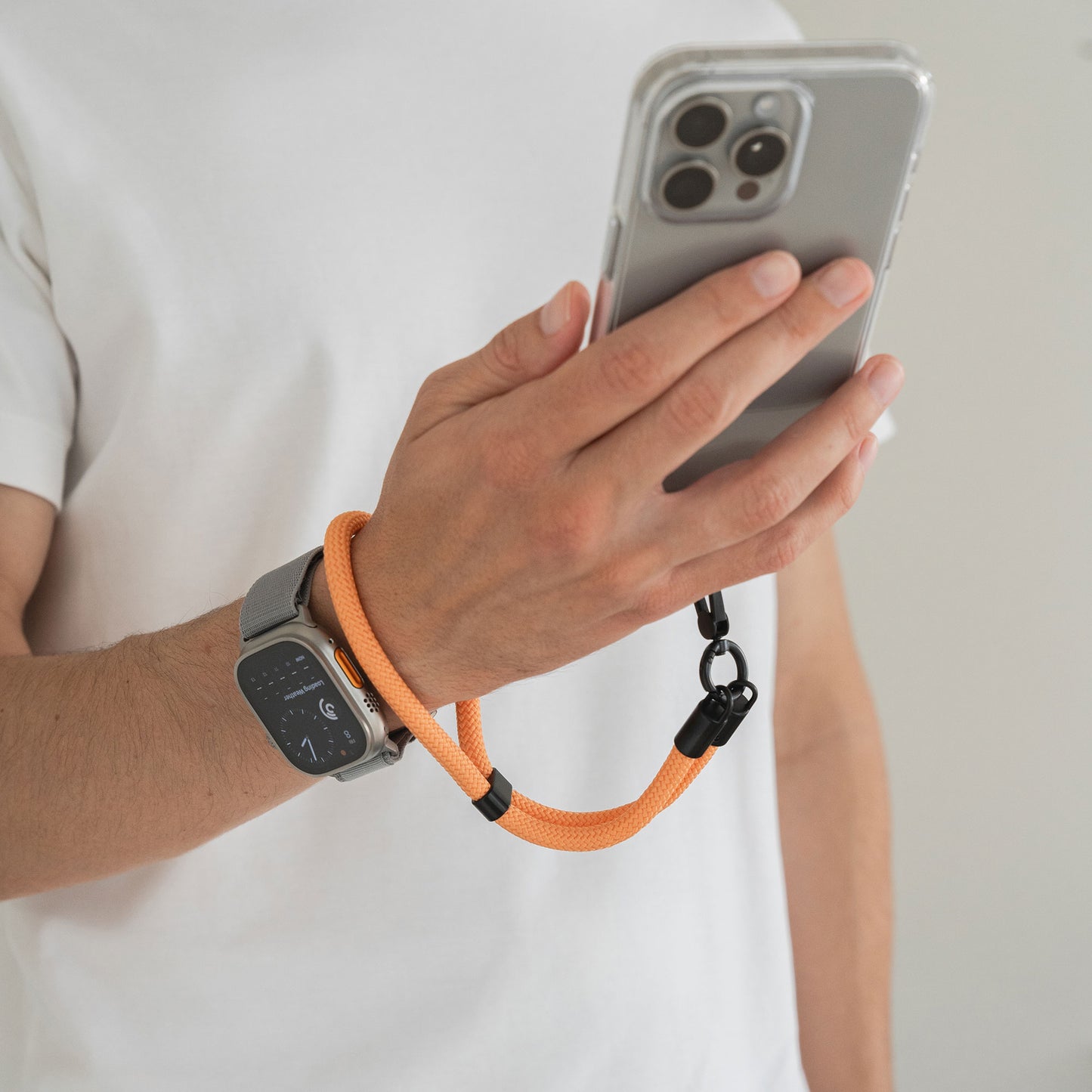 Eine Person hält ein Smartphone mit der orangefarbenen arktis Lanyard Handykette fürs Handgelenk von Arktis, trägt eine Smartwatch und ein weißes T-Shirt – und zeigt, dass Arktis Smartphone Zubehör auch ein stilvolles Mode-Accessoire ist.