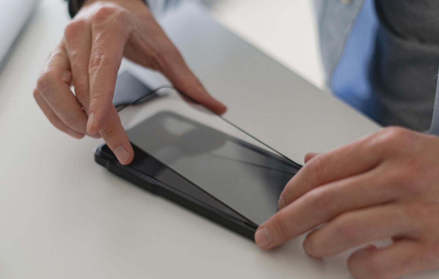 Eine Person richtet das ArktisPRO iPhone 15 Pro Max FULL COVER Displayschutz GLAS (hüllenfreundlich) mit beiden Händen auf einem flach auf einer weißen Oberfläche liegenden Smartphone aus, wobei das kratzfeste Glas das Telefon teilweise bedeckt.