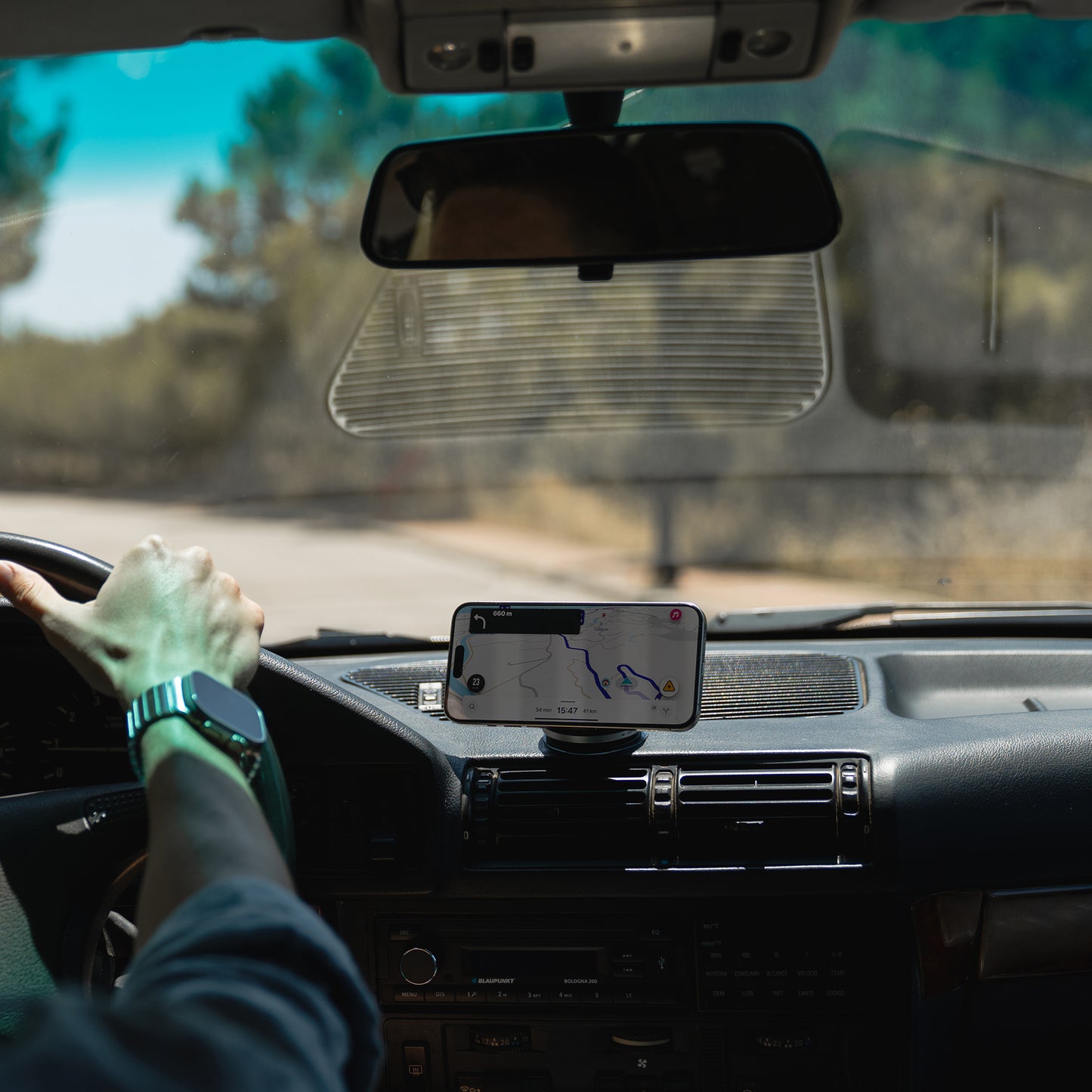 Die Hand des Fahrers am Lenkrad, das iPhone mit GPS-Navigation gesichert an einer arktis MagicDock iPhone MagSafe Halterung von arktis auf dem Armaturenbrett, Straße und Bäume voraus.
