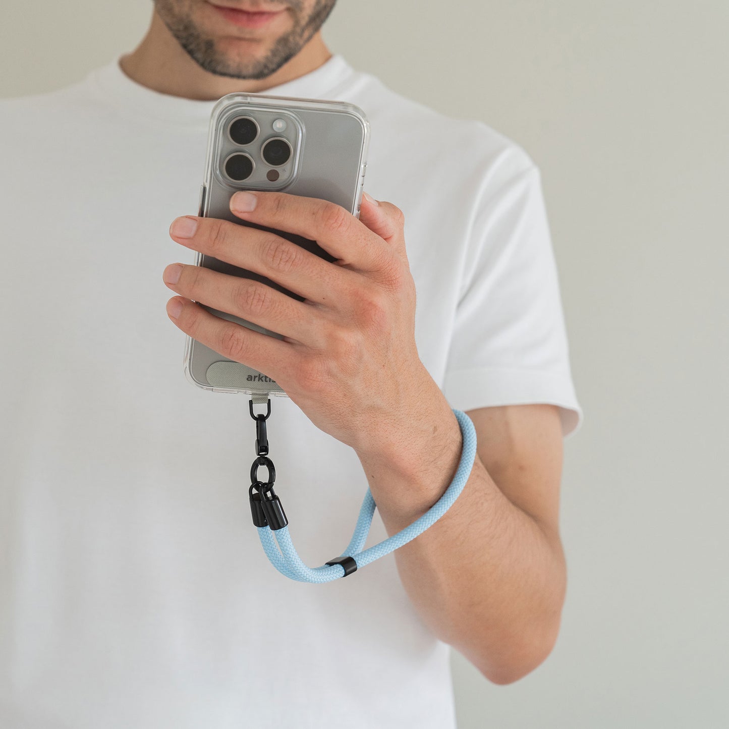 Ein Mann in einem weißen Hemd hält ein Telefon mit der daran befestigten Arktis Lanyard Handykette fürs Handgelenk von Arktis und präsentiert dieses trendige Smartphone-Zubehör mit verstellbarer Länge.