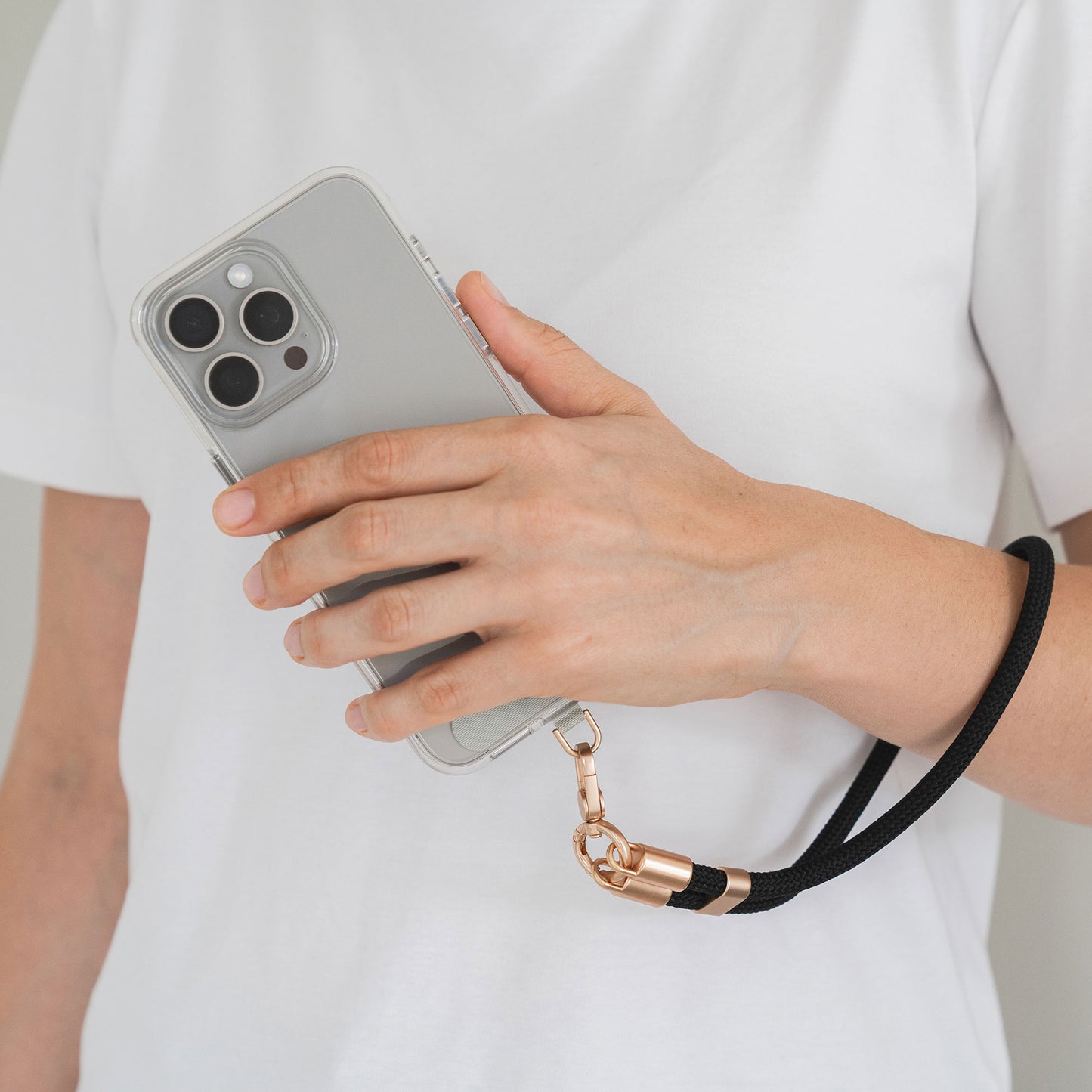 Eine Person in einem weißen T-Shirt hält ein Smartphone mit einer Arktis Lanyard Handykette fürs Handgelenk von Arktis, mit einem durchsichtigen Gehäuse und einem schwarzen Armband mit goldenem Anschluss – ein stilvolles Smartphone-Accessoire.