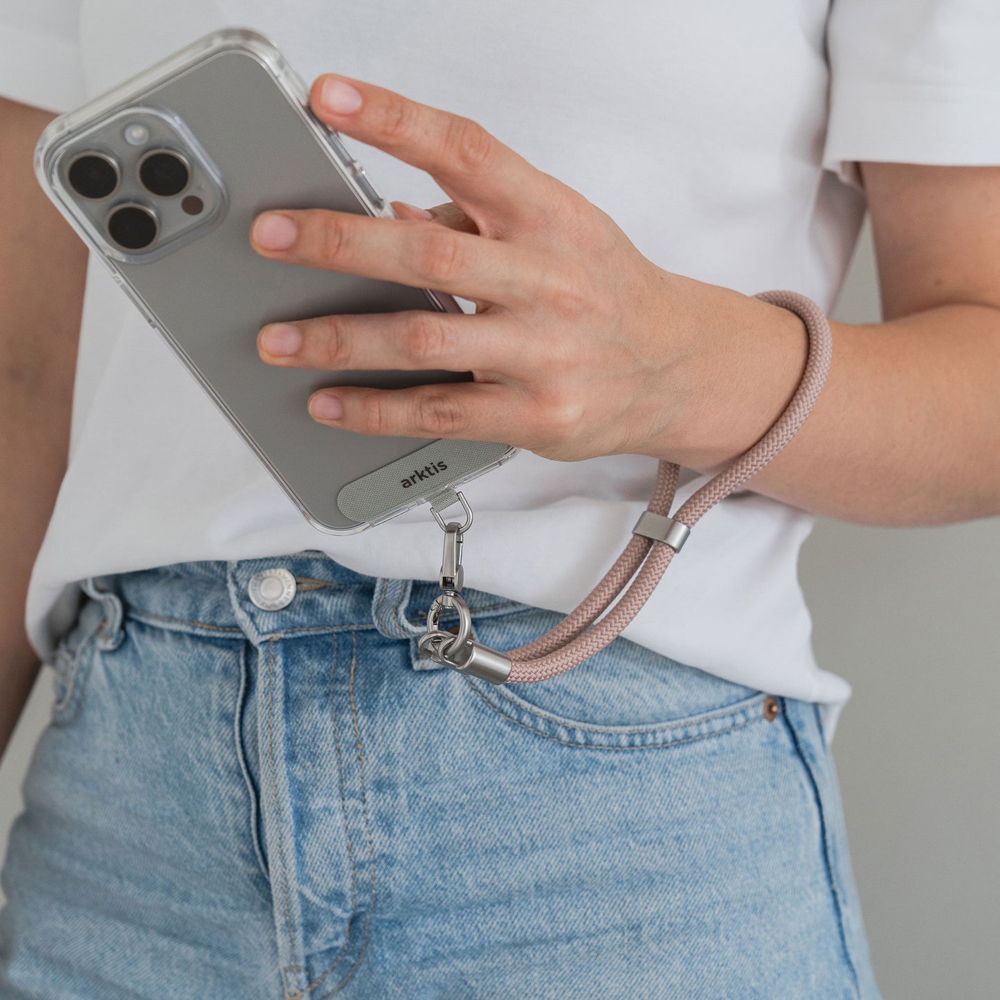 Eine Person hält ein Telefon mit einer durchsichtigen Hülle und einer rosafarbenen Arktis-Lanyard-Handykette fürs Handgelenk von Arktis und trägt ein weißes Hemd und blaue Jeans – ein ideales Mode-Accessoire für Fans von stylischem Smartphone-Zubehör.