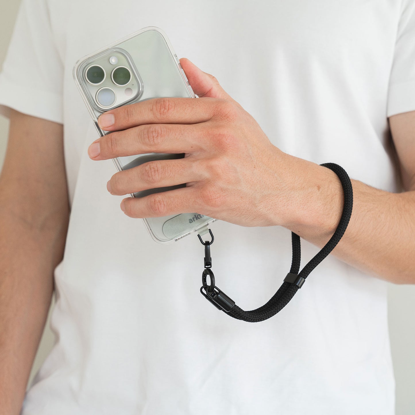 In der Hand hält man ein Smartphone in einer durchsichtigen Hülle, an der die schwarze Arktis Lanyard Handykette fürs Handgelenk (verstellbare Länge) befestigt ist, und trägt es über einem weißen Hemd – Ihr stilvolles, bequemes Smartphone-Accessoire von Arktis.