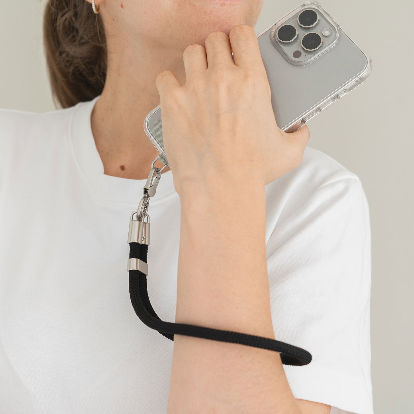 Eine Person in einem klassischen weißen T-Shirt hält ein Smartphone mit der Arktis Lanyard Handykette fürs Handgelenk von Arktis – einer durchsichtigen Hülle mit schwarzem Armband für Stil und Komfort.