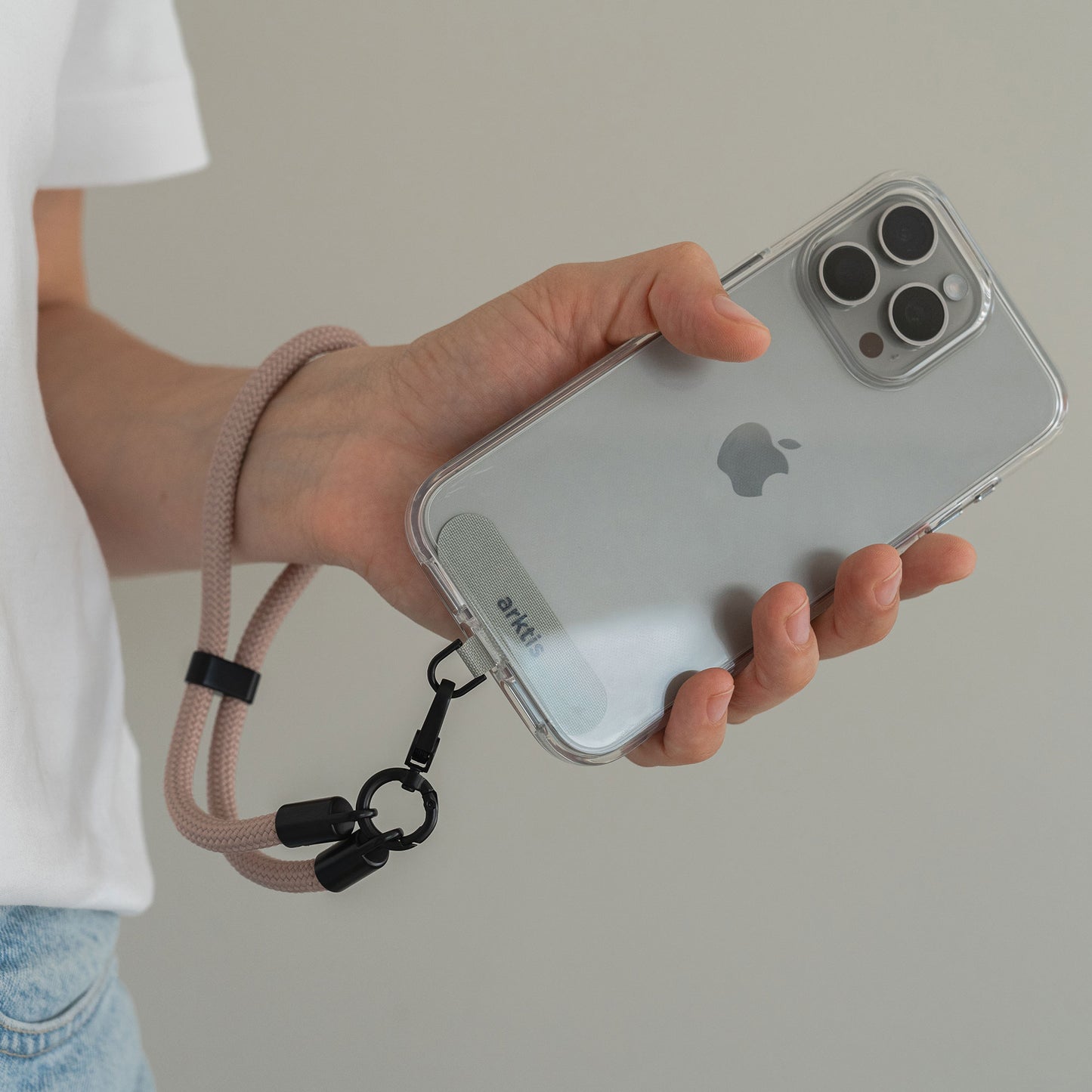 Eine Hand hält ein iPhone mit einer durchsichtigen Hülle und die Arktis Lanyard Handykette fürs Handgelenk von Arktis, ein stilvolles Smartphone-Zubehör mit einstellbarer Länge für mehr Komfort.