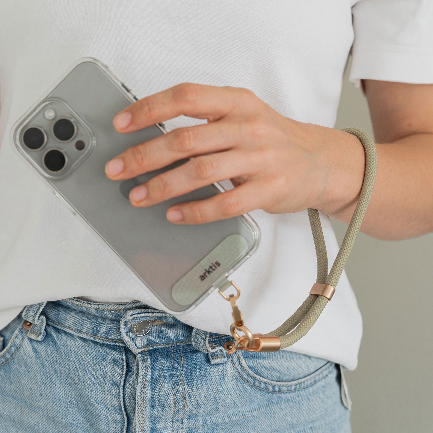 Eine Person in einem weißen T-Shirt und blauen Jeans hält ein Smartphone mit der Arktis Lanyard Handykette fürs Handgelenk von Arktis an ihrer Hüfte befestigt. Die beigefarbene Handschlaufe ermöglicht eine sichere und praktische Nutzung des Smartphone-Accessoires.