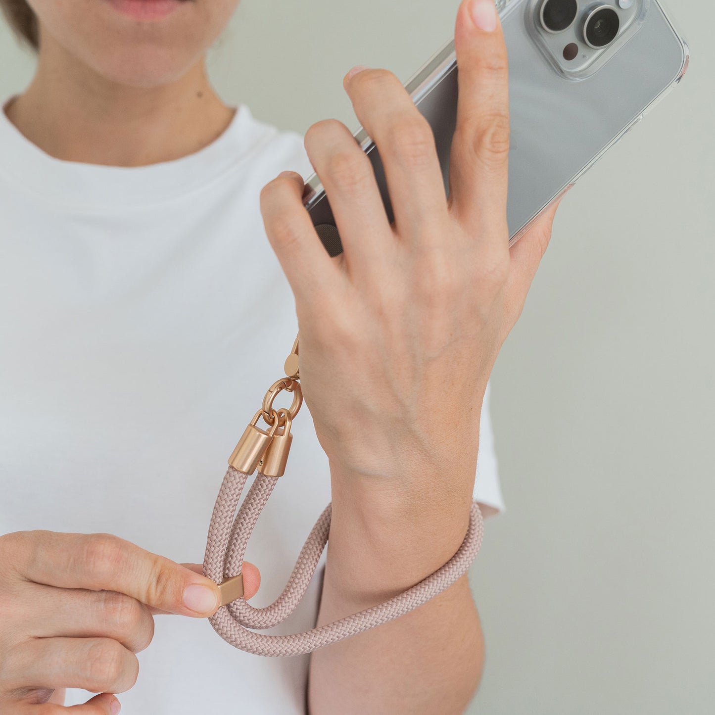 Eine Person im weißen Hemd hält ein Smartphone mit der arktis Lanyard Handykette fürs Handgelenk von Arktis, einem rosa geflochtenen Smartphone-Zubehör, um ihr Handgelenk geschlungen und zwischen ihren Fingern gehalten.
