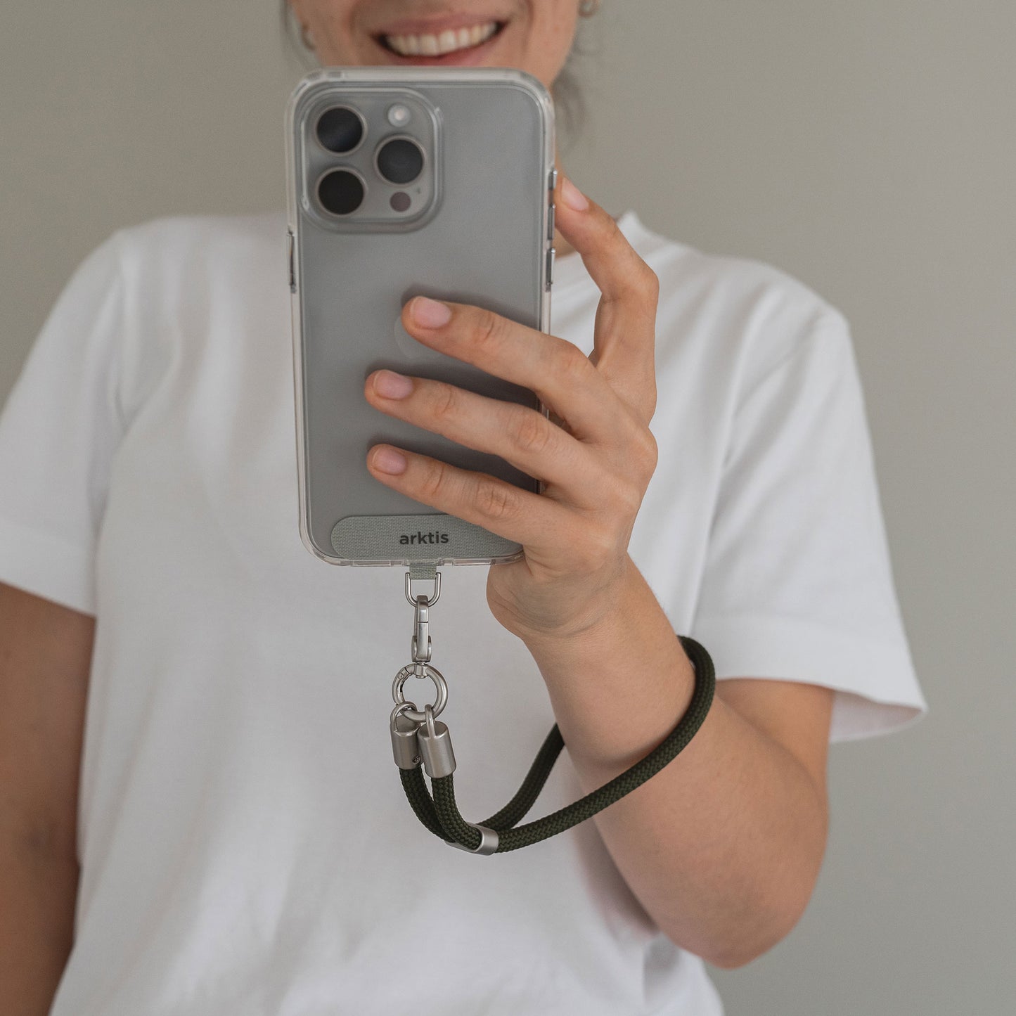 Eine Person im weißen Hemd hält ein Telefon mit der Arktis Lanyard Handykette fürs Handgelenk und lächelt in die Kamera – das ideale Smartphone-Zubehör von Arktis für alle, die Wert auf Stil und Funktionalität legen.
