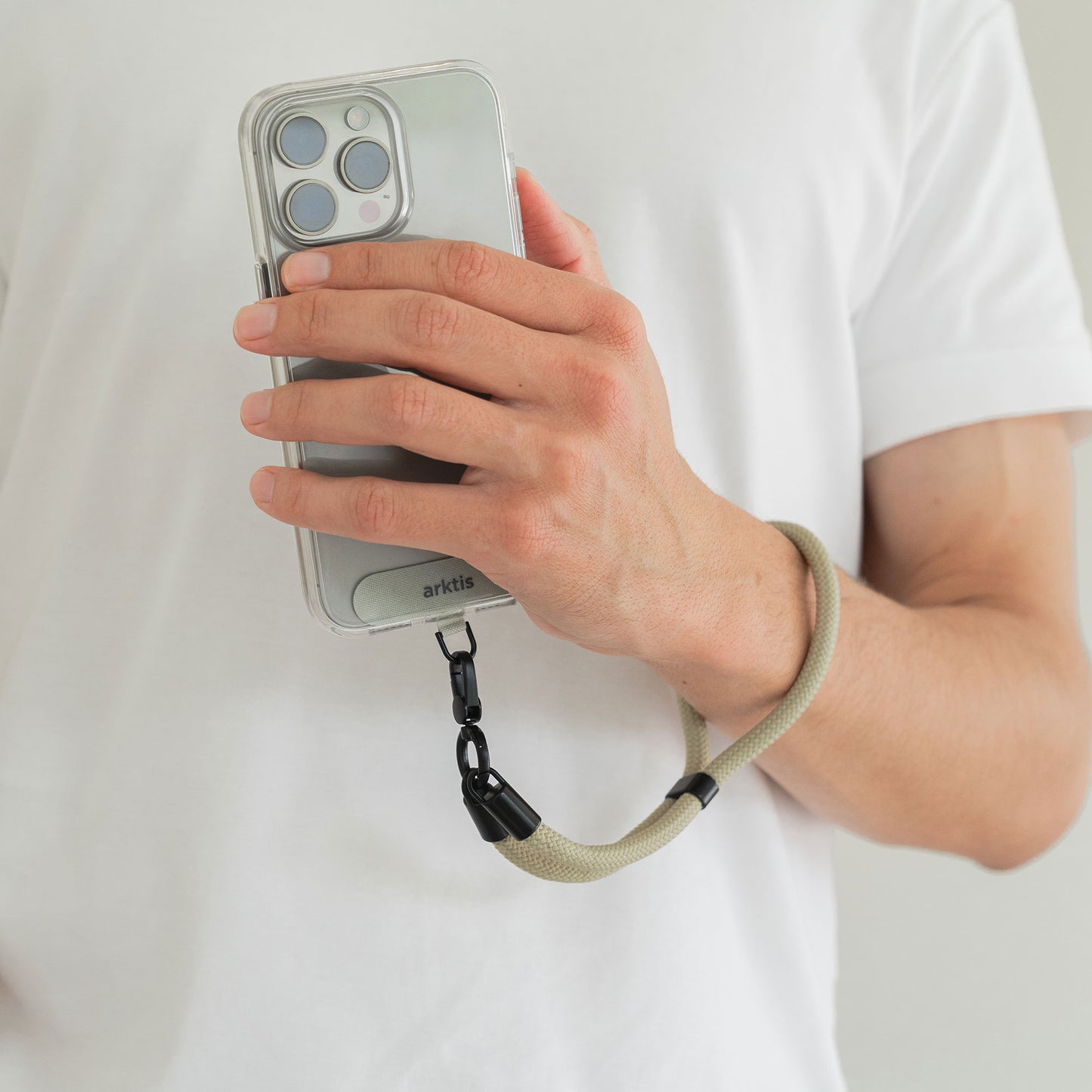 Eine Person in einem weißen Hemd hält ein Telefon mit der Arktis Lanyard Handykette fürs Handgelenk von Arktis, die über ein durchsichtiges Gehäuse und ein beiges, längenverstellbares Schlüsselband als praktisches Smartphone-Zubehör verfügt.