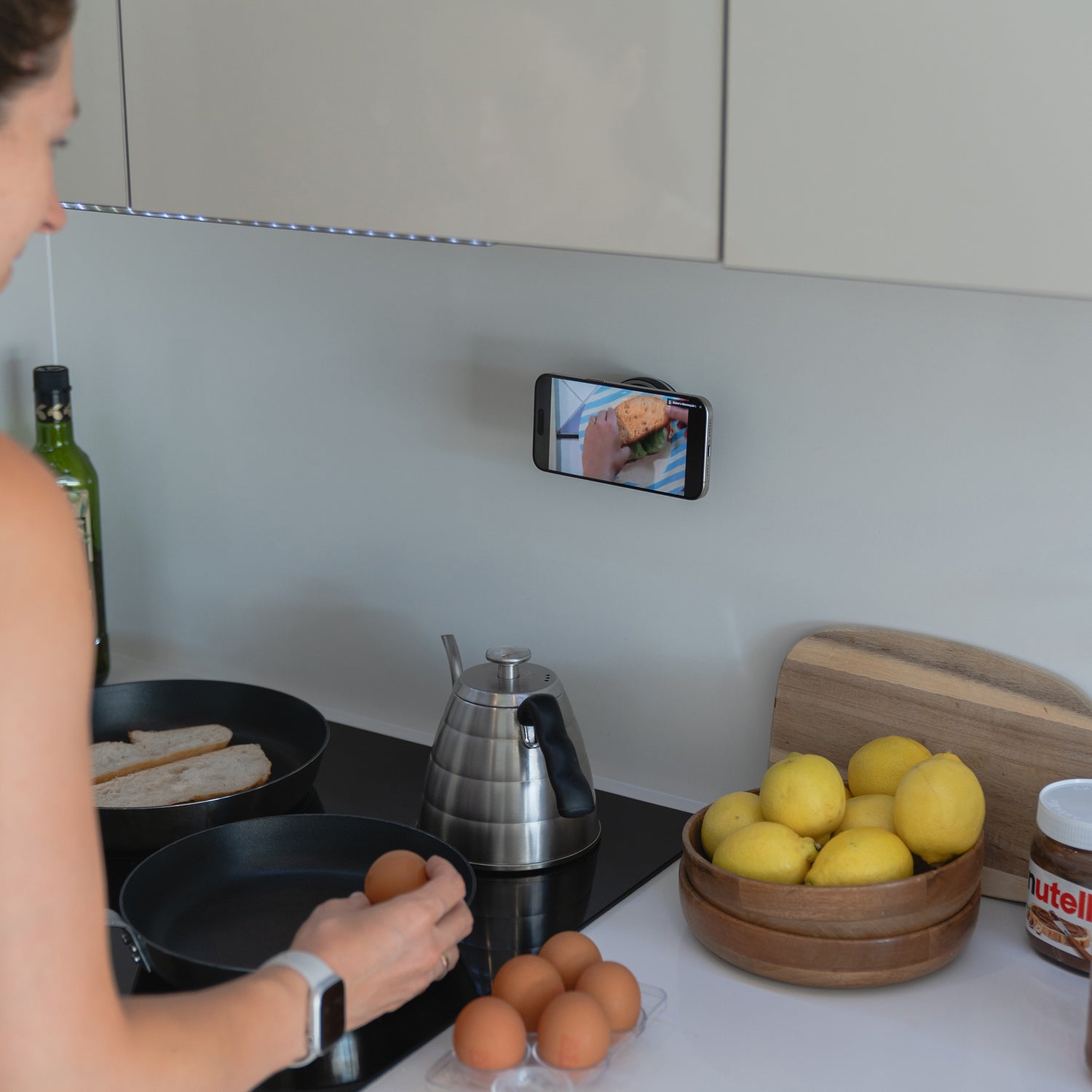 Eine Person in der Küche bereitet Eier und Toast zu, während sie ein Kochvideo auf ihrem Handy anschaut, das mit der eleganten arktis MagicDock iPhone MagSafe Halterung sicher an der Wand befestigt ist.