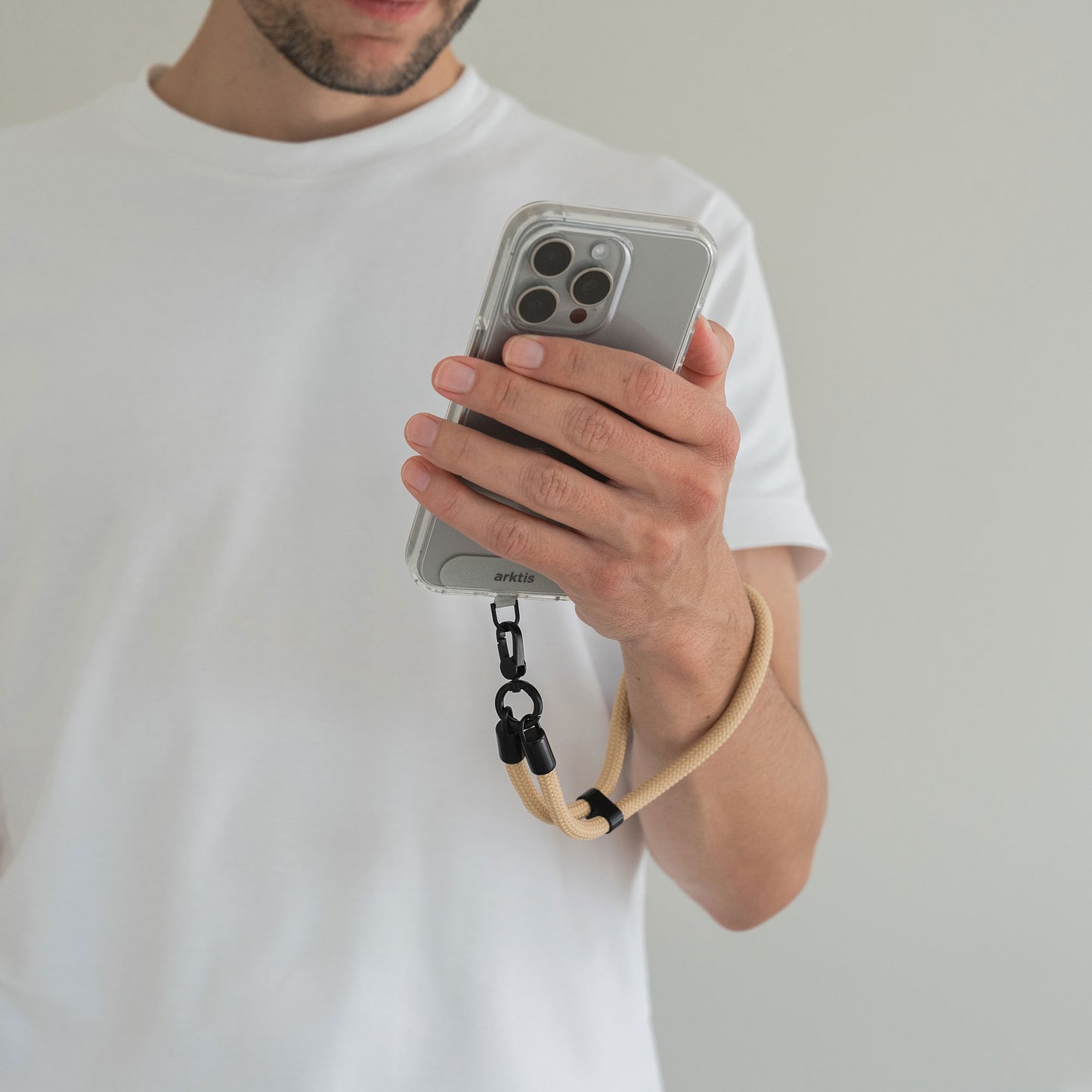 Ein Mann in einem weißen T-Shirt hält ein Telefon, an dem die Arktis Lanyard Handykette fürs Handgelenk von Arktis befestigt ist, ein stilvolles Smartphone-Accessoire mit verstellbarer Länge für zusätzlichen Komfort.